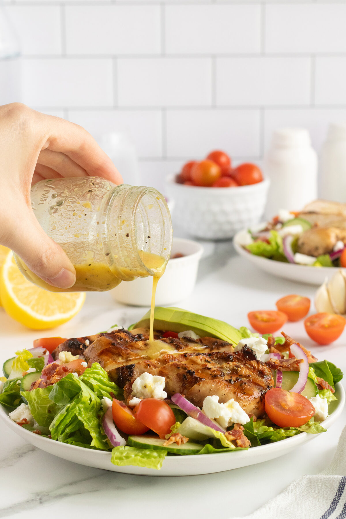 A hand pours vinaigrette from a jar onto a salad with grilled chicken, cherry tomatoes, red onion, avocado, and feta on a white plate. In the background are more vegetables and a half lemon on a white countertop.