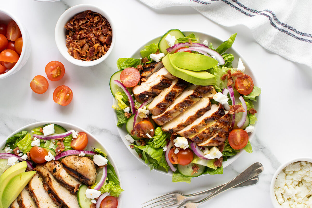 A plate of grilled chicken salad with sliced avocado, cherry tomatoes, red onions, bacon bits, and crumbled cheese on lettuce, with extra salad, tomatoes, and bacon bits in small bowls nearby, on a white marble surface.