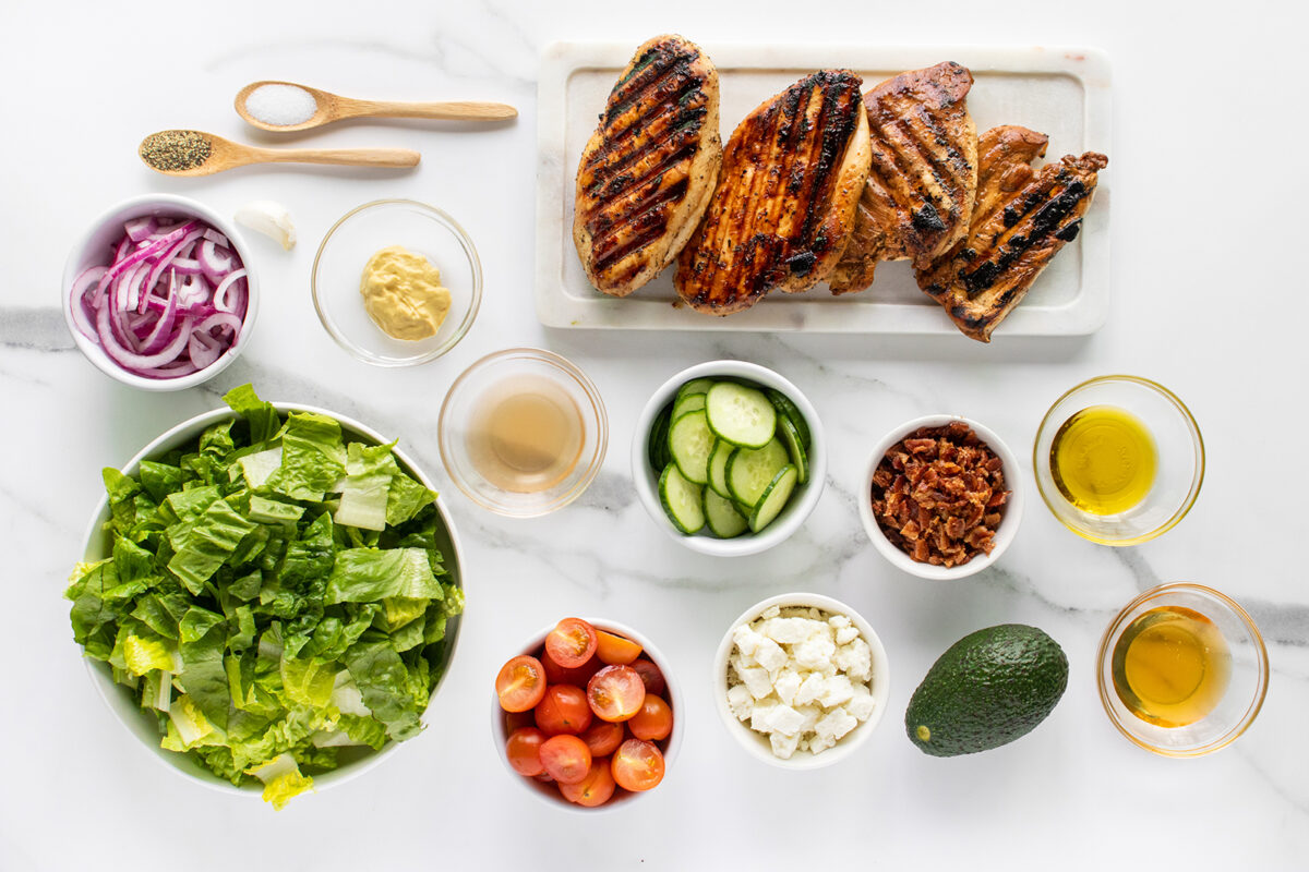 A variety of fresh salad ingredients and grilled chicken breasts are neatly arranged on a white surface. Ingredients include lettuce, cherry tomatoes, cucumber, red onion, avocado, bacon, feta, seasonings, olive oil, vinegar, and mustard.