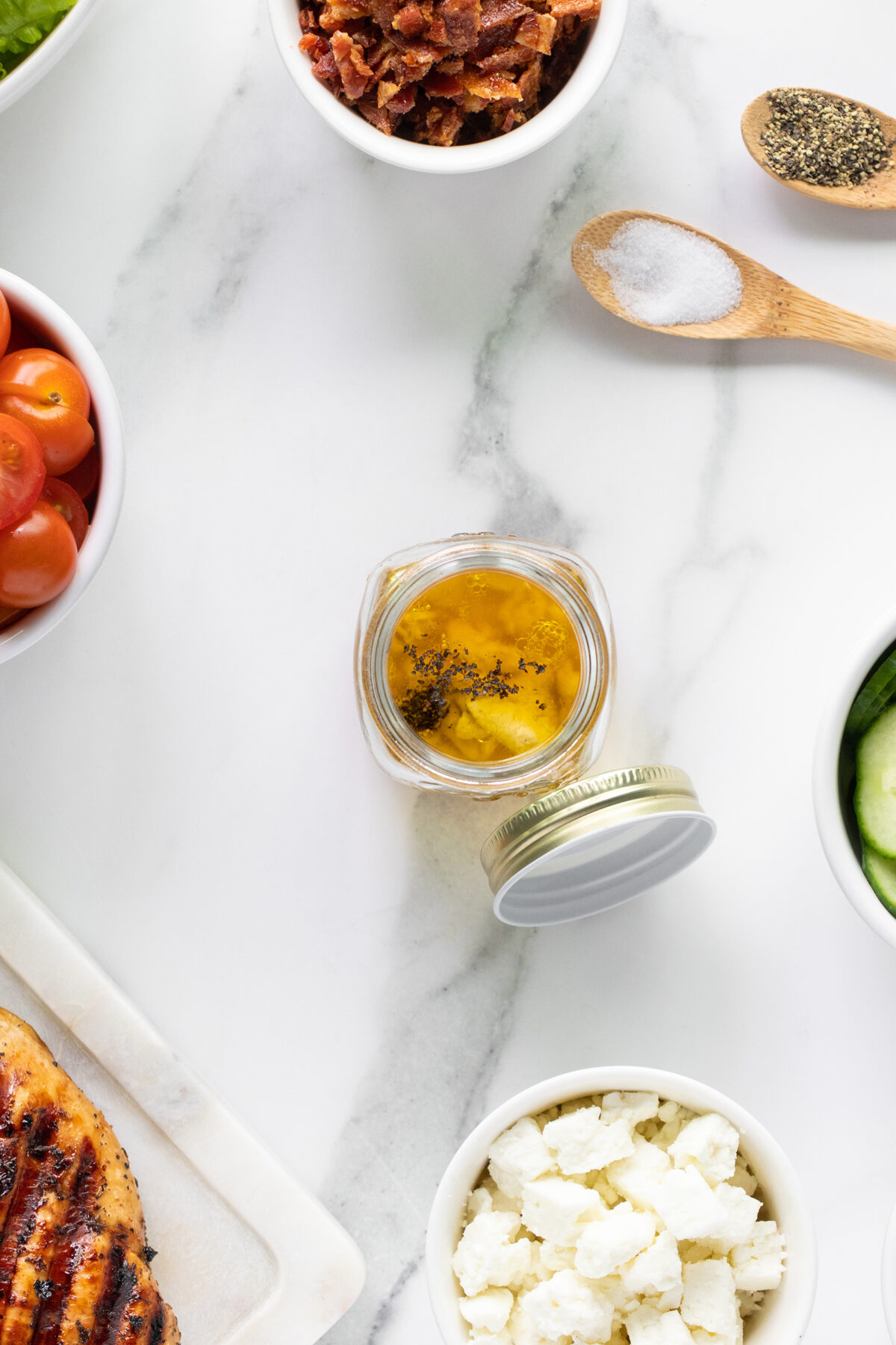 Overhead view of a jar of salad dressing surrounded by small bowls of cherry tomatoes, bacon bits, cucumber slices, crumbled cheese, salt, pepper, and part of a grilled chicken breast on a white marble surface.