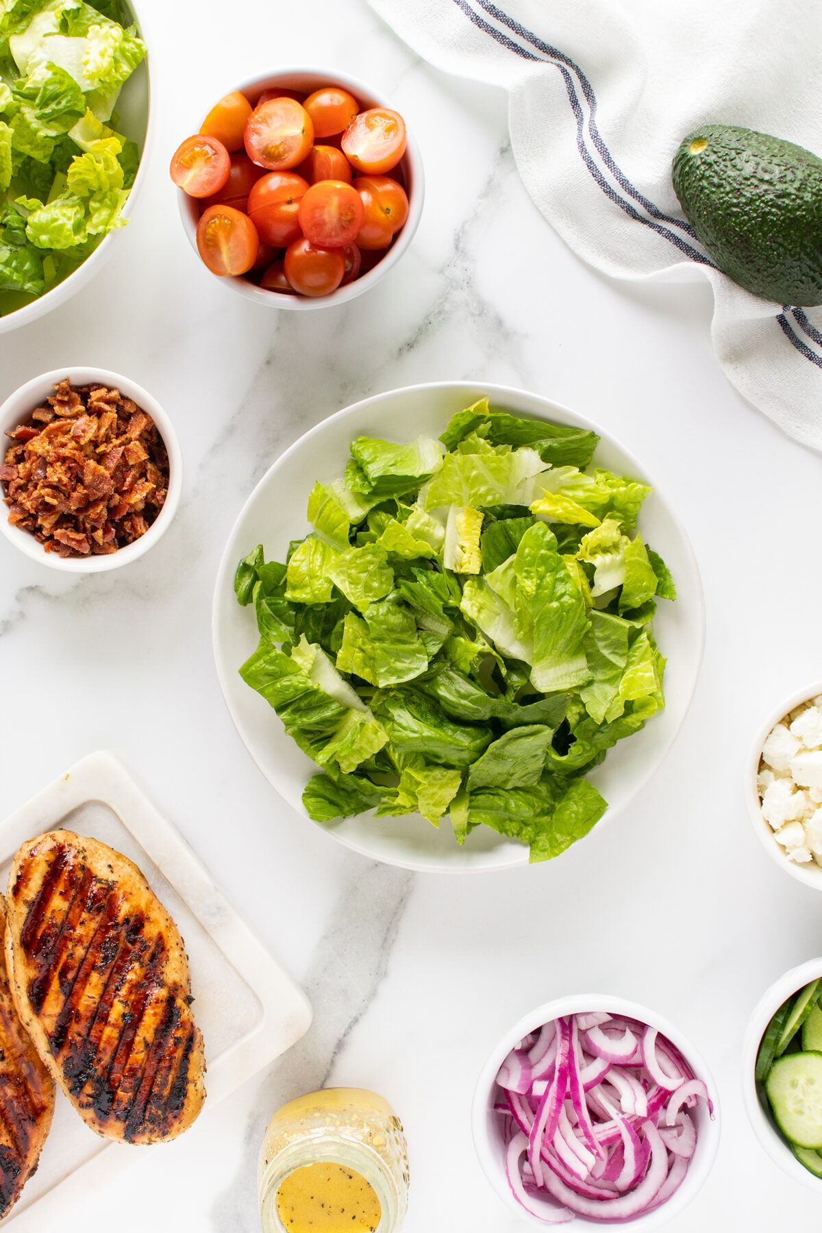 A flat lay of salad ingredients on a white surface, including chopped lettuce, cherry tomatoes, avocado, bacon bits, grilled chicken, sliced red onions, cucumber, crumbled cheese, and a jar of dressing.