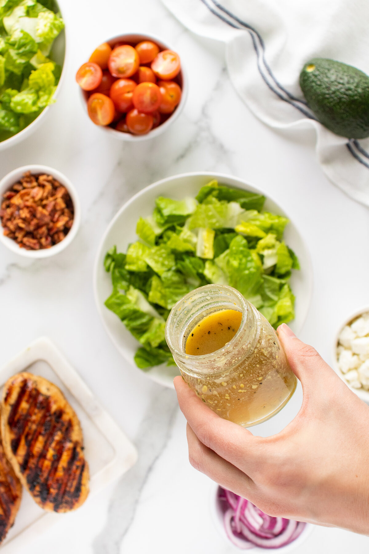 A hand holds a jar of vinaigrette dressing above a plate of chopped romaine lettuce, surrounded by small bowls of cherry tomatoes, bacon bits, avocado, grilled chicken, feta cheese, and red onions.