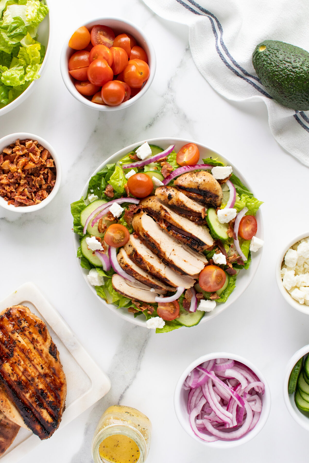 A bowl of salad topped with sliced grilled chicken, cherry tomatoes, red onions, feta cheese, and bacon bits, surrounded by small bowls of vegetables, cheese, and a grilled chicken breast on a marble surface.