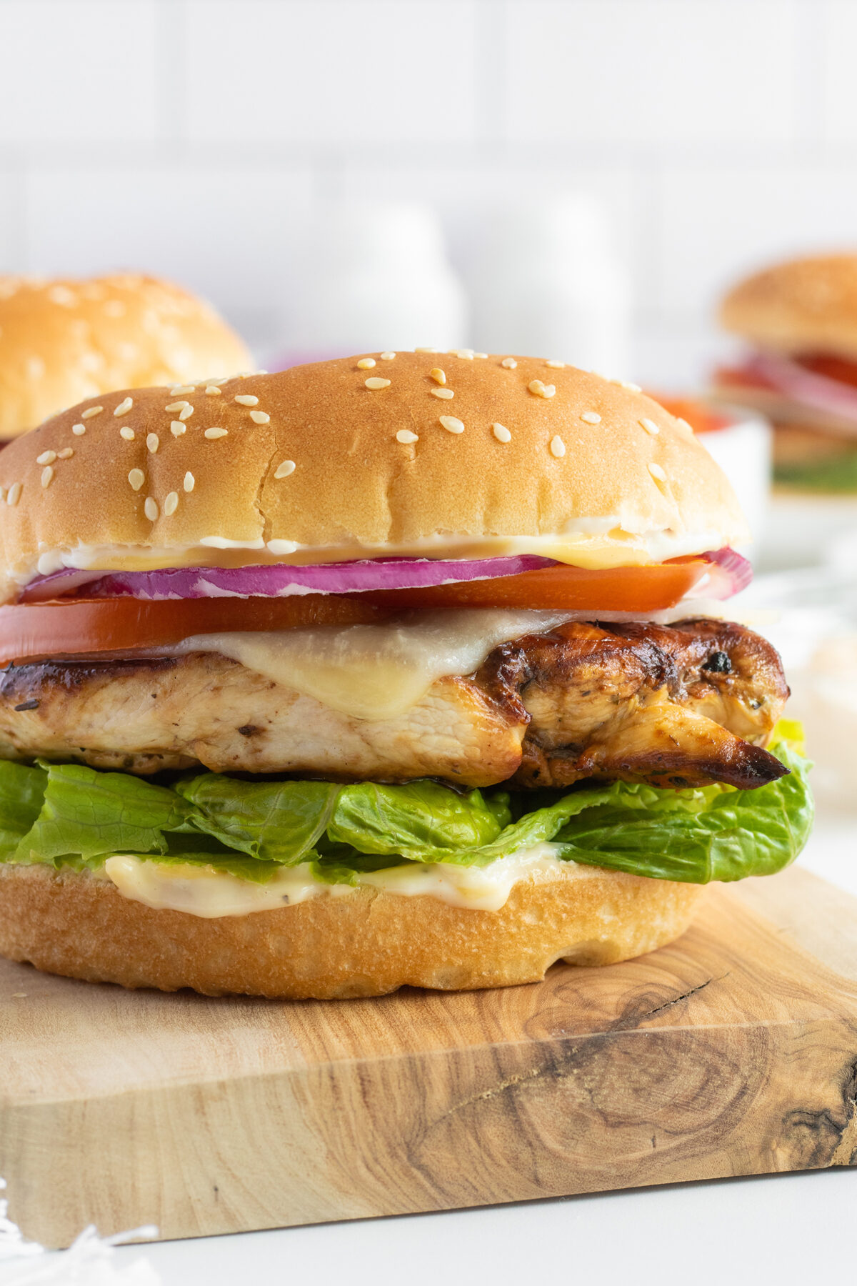 A grilled chicken sandwich with lettuce, melted cheese, tomato, and red onion on a sesame seed bun, placed on a wooden board.