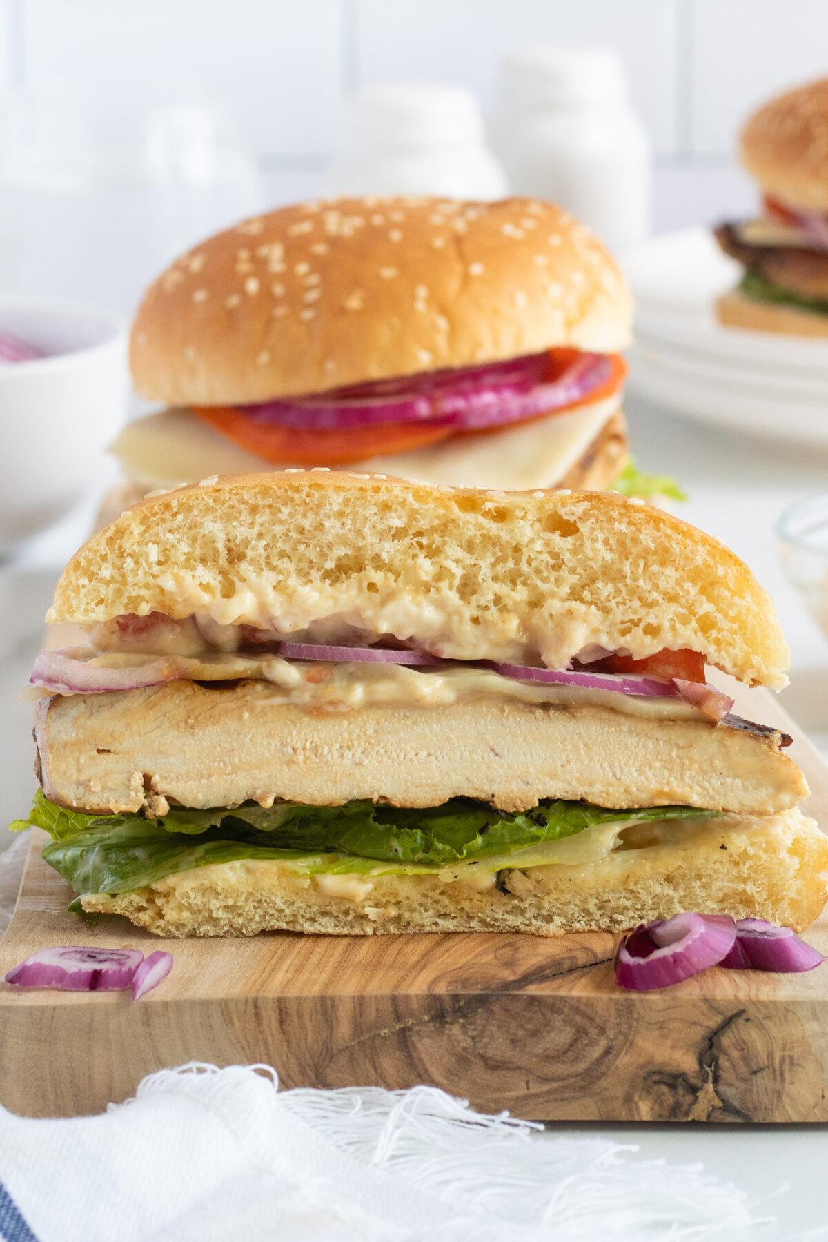 A close-up of a sliced chicken sandwich on a wooden board, with lettuce, tomato, red onion, and sauce in a sesame bun. Another sandwich and ingredients are blurred in the background.