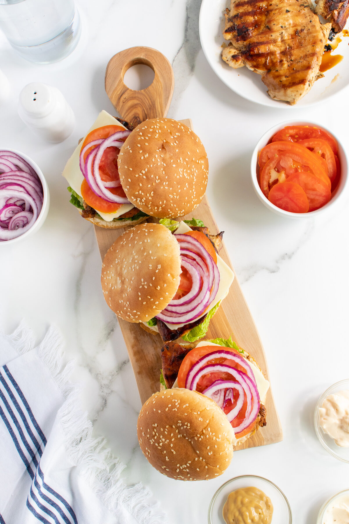 Three chicken sandwiches with cheese, lettuce, tomato, and red onion on a wooden board, surrounded by sliced onions, tomatoes, grilled chicken breast, mustard, and a glass of water on a white table.