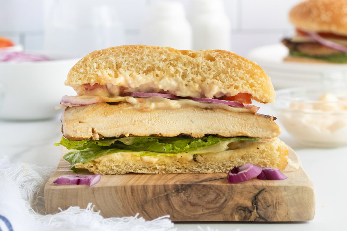 A grilled chicken sandwich on a wooden board, cut in half to show layers of chicken, lettuce, tomato, red onion, and sauce on a bun. Some red onion pieces are scattered on the board.