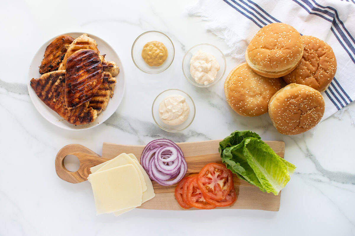 Grilled chicken breasts, burger buns, mustard, mayonnaise, cheese slices, red onion rings, tomato slices, and romaine lettuce arranged on a table, ready for making chicken sandwiches.