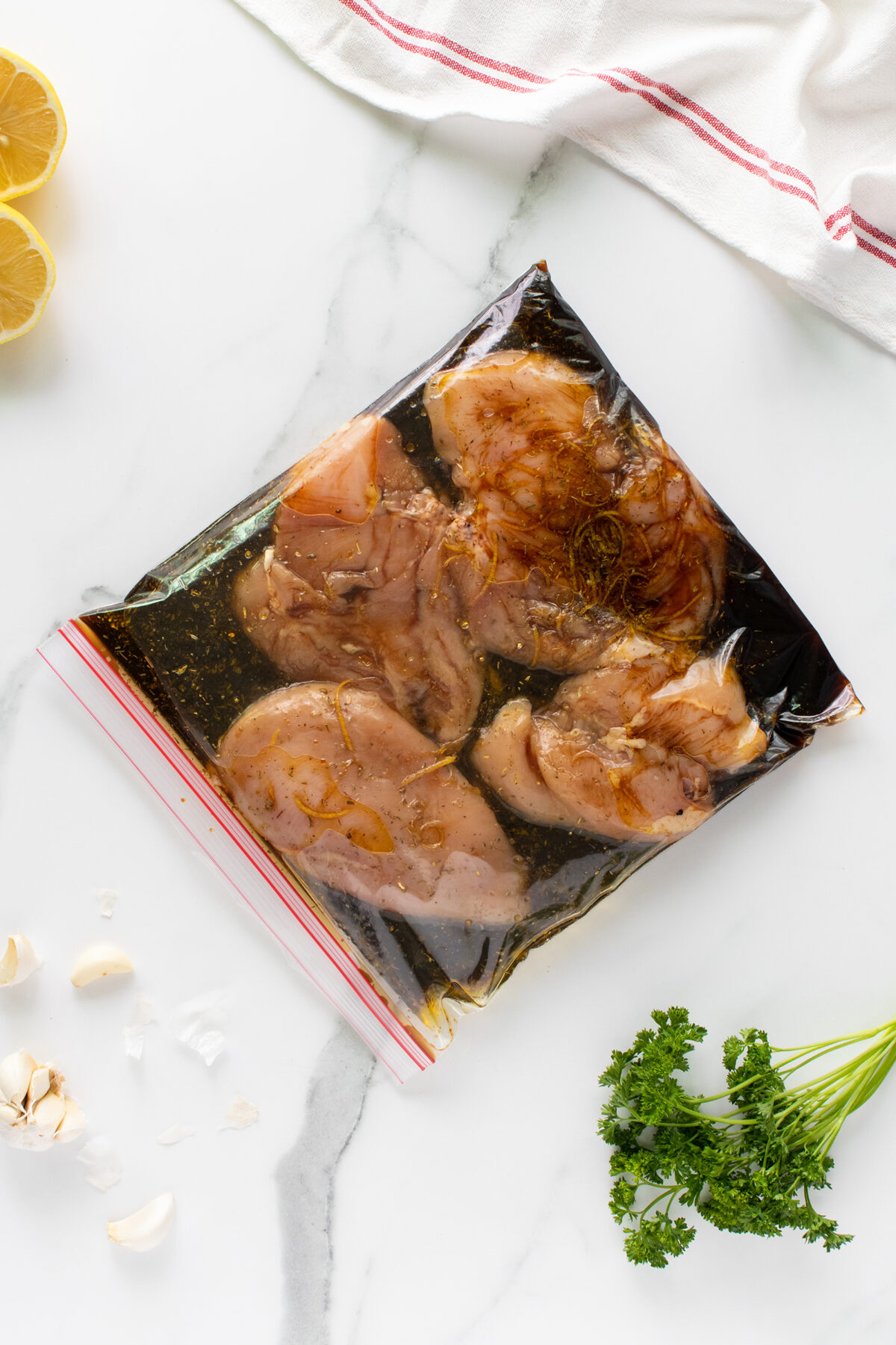 Raw chicken breasts marinating in a clear ziplock bag with herbs and spices, placed on a white marble surface. Surrounding are parsley, garlic cloves, lemon halves, and a white towel with red stripes.