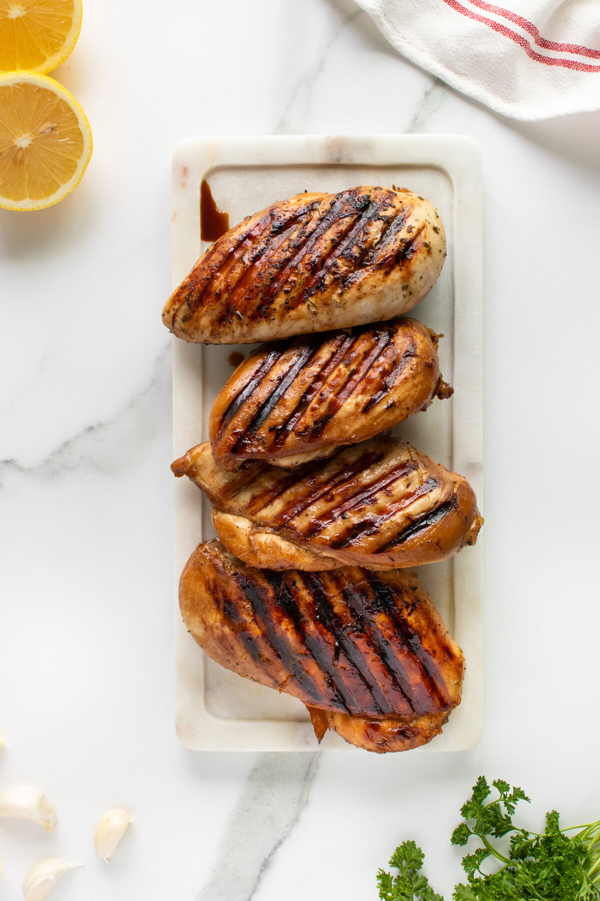 Four grilled chicken breasts with char marks are arranged on a white marble platter. Lemon halves, parsley, garlic cloves, and a red-striped towel are nearby on the marble surface.