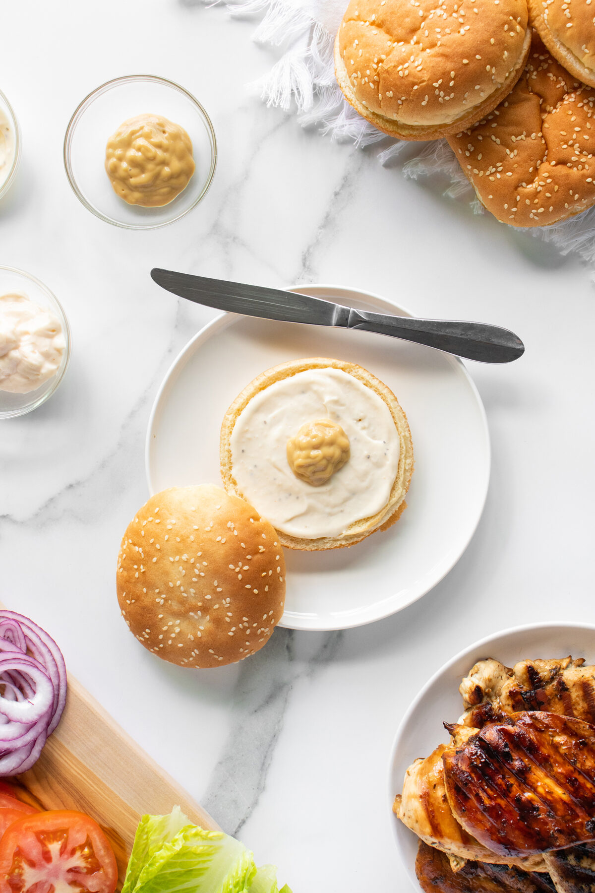 Open hamburger bun with mayonnaise and mustard on a white plate, a knife resting on top, surrounded by buns, grilled chicken, sliced onions, tomatoes, lettuce, and condiments on a marble surface.
