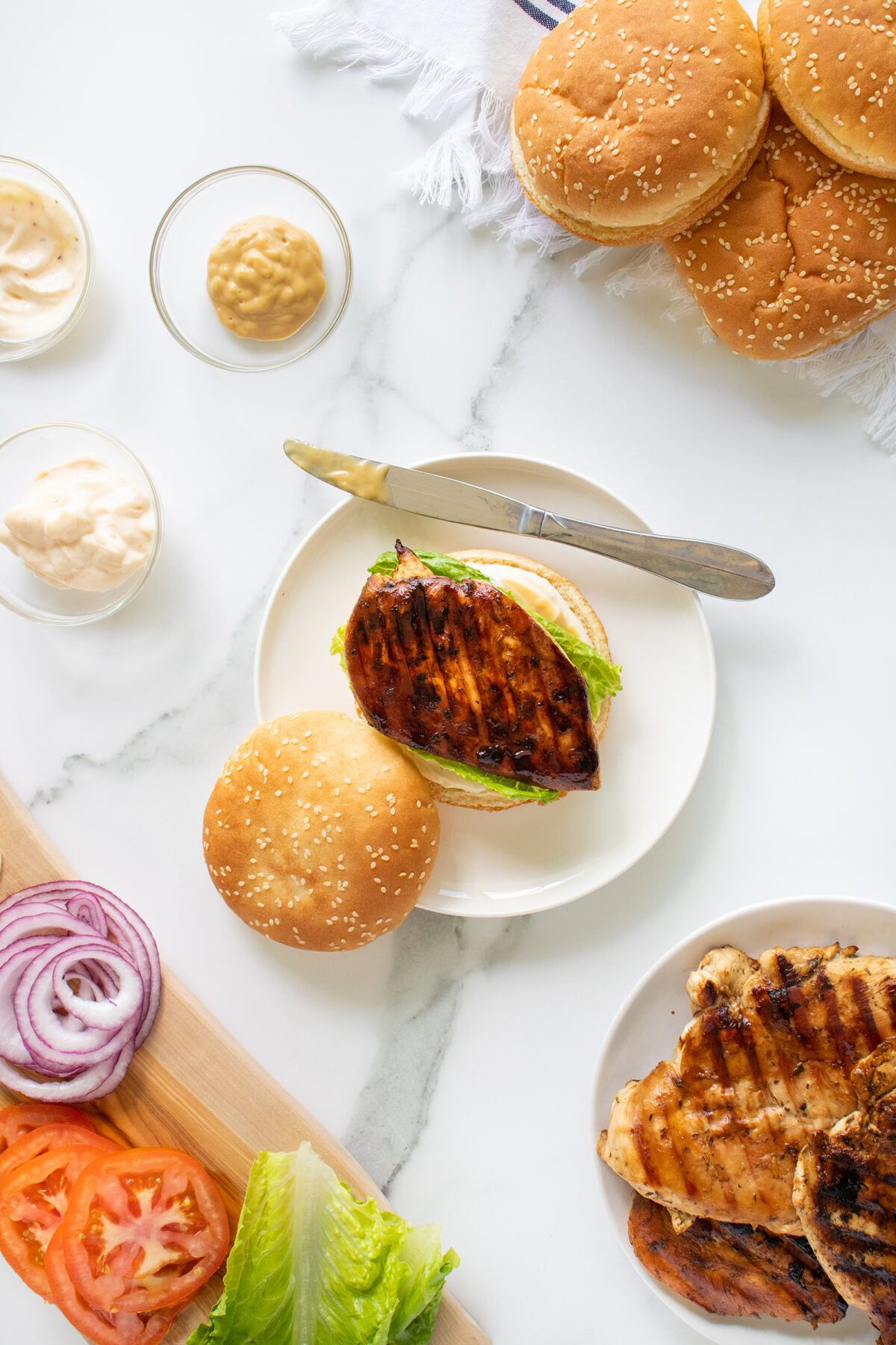 A grilled chicken sandwich with lettuce on a sesame bun sits on a plate with a knife, surrounded by extra buns, grilled chicken, sliced tomatoes, red onions, lettuce, and small bowls of mayo and mustard on a white surface.