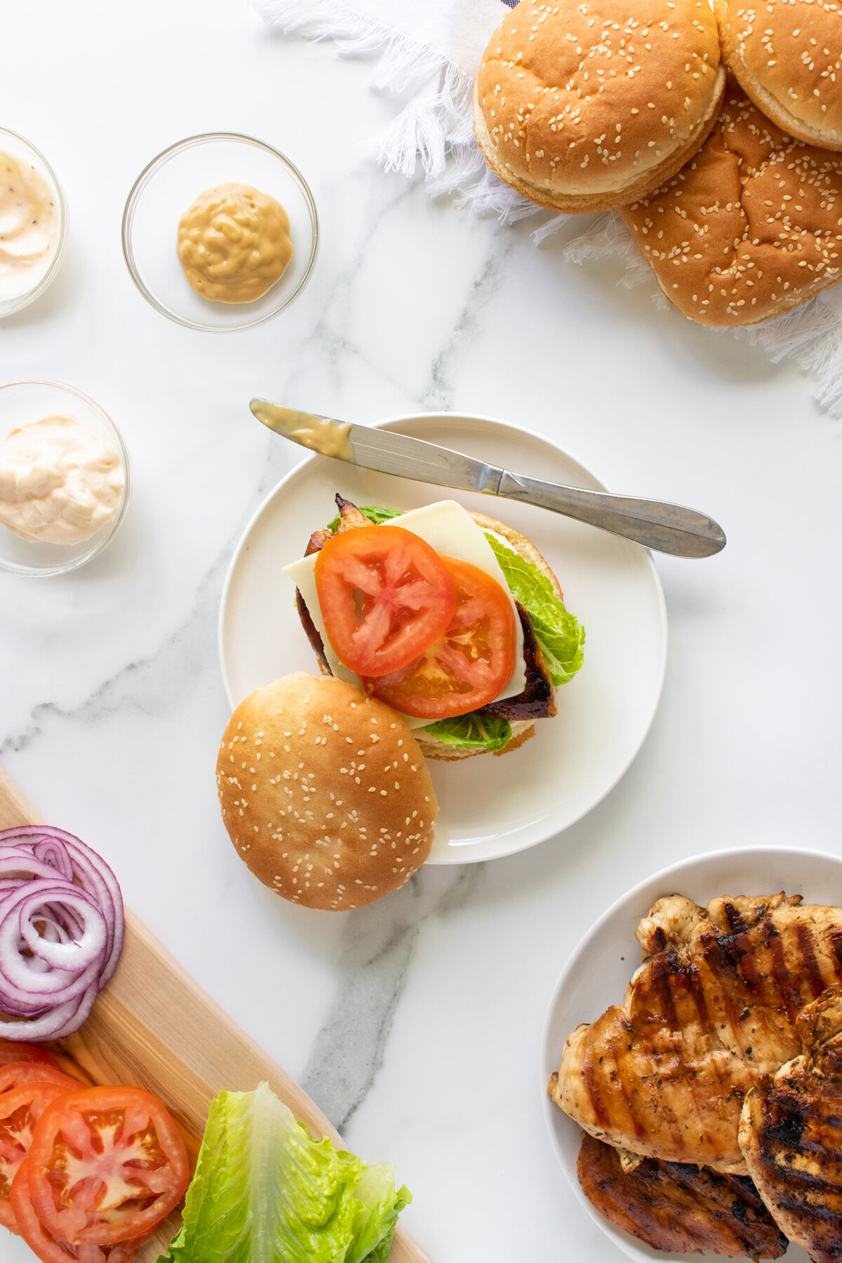 Top-down view of a chicken sandwich with tomato, lettuce, and cheese on a sesame bun, surrounded by grilled chicken, sliced vegetables, sauces, and extra buns on a white surface.
