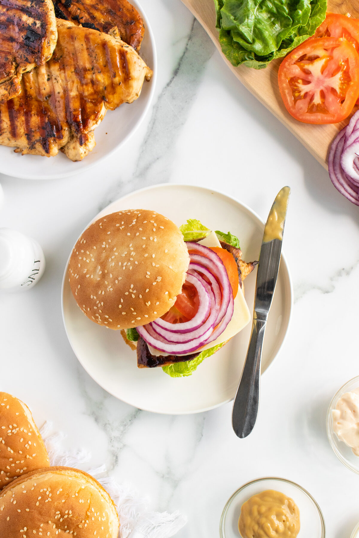 A plate with a sandwich bun, lettuce, cheese, grilled chicken, red onion, and tomato. A knife with mustard rests on the plate. Surrounding are grilled chicken, sliced lettuce, tomato, red onion, buns, and condiments.