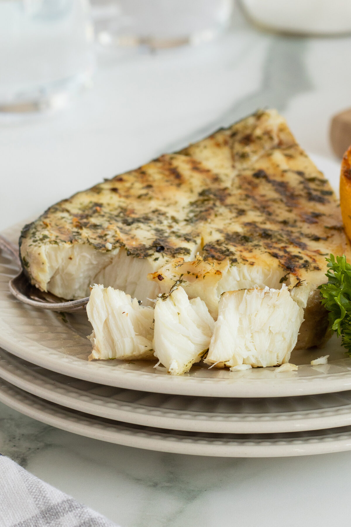 A grilled fish steak with herbs sits on a plate, with a piece flaked off by a fork, garnished with parsley and a lemon wedge. The plate rests on a stack of dishes on a light surface.