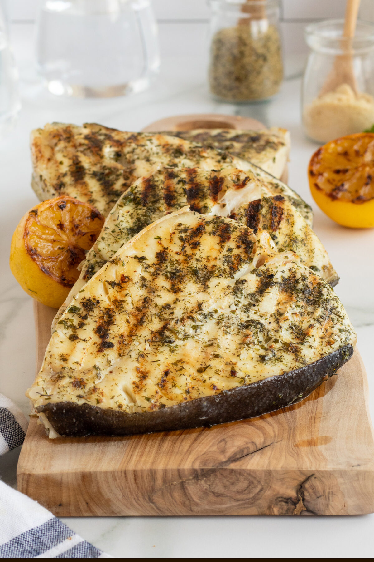 Three grilled fish steaks with herbs rest on a wooden cutting board, accompanied by two grilled lemon halves. Jars of spices and a checkered cloth are visible in the background.