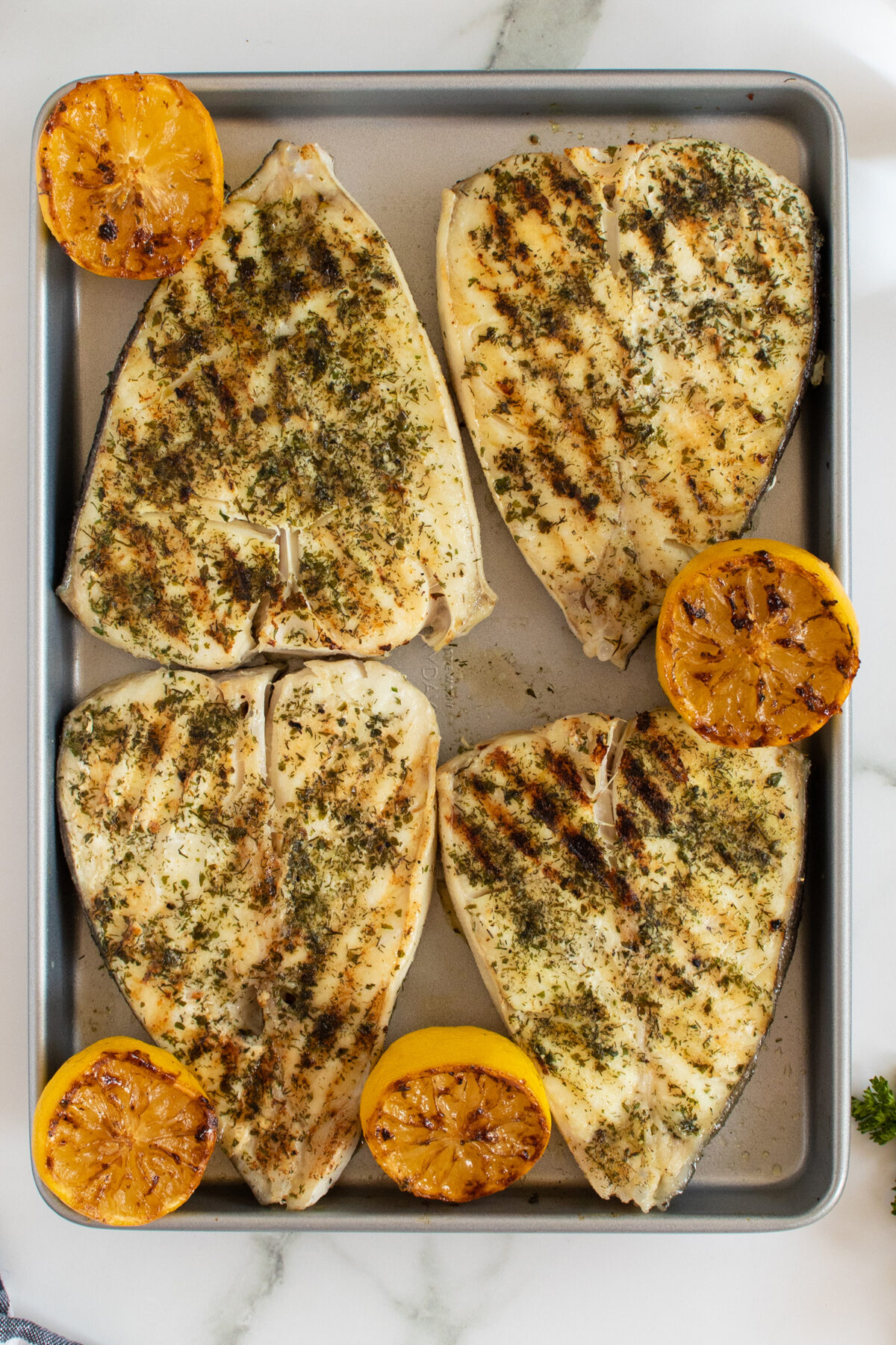 Four grilled fish fillets, seasoned with herbs, are arranged on a baking sheet with four halves of grilled lemon. The surface is lightly charred, and the background includes a white marble countertop.