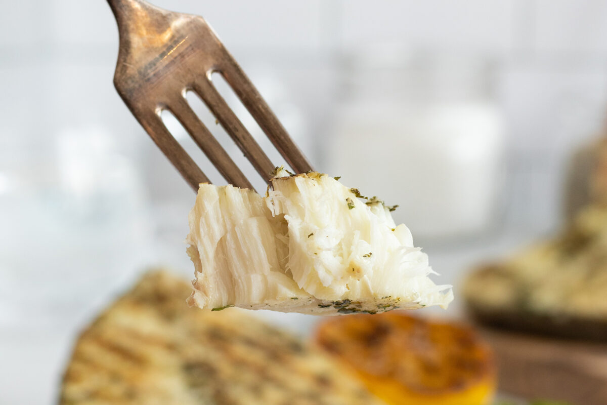 A close-up of a fork holding a flaky, seasoned piece of white fish, with a blurred background featuring grilled food and a slice of lemon.