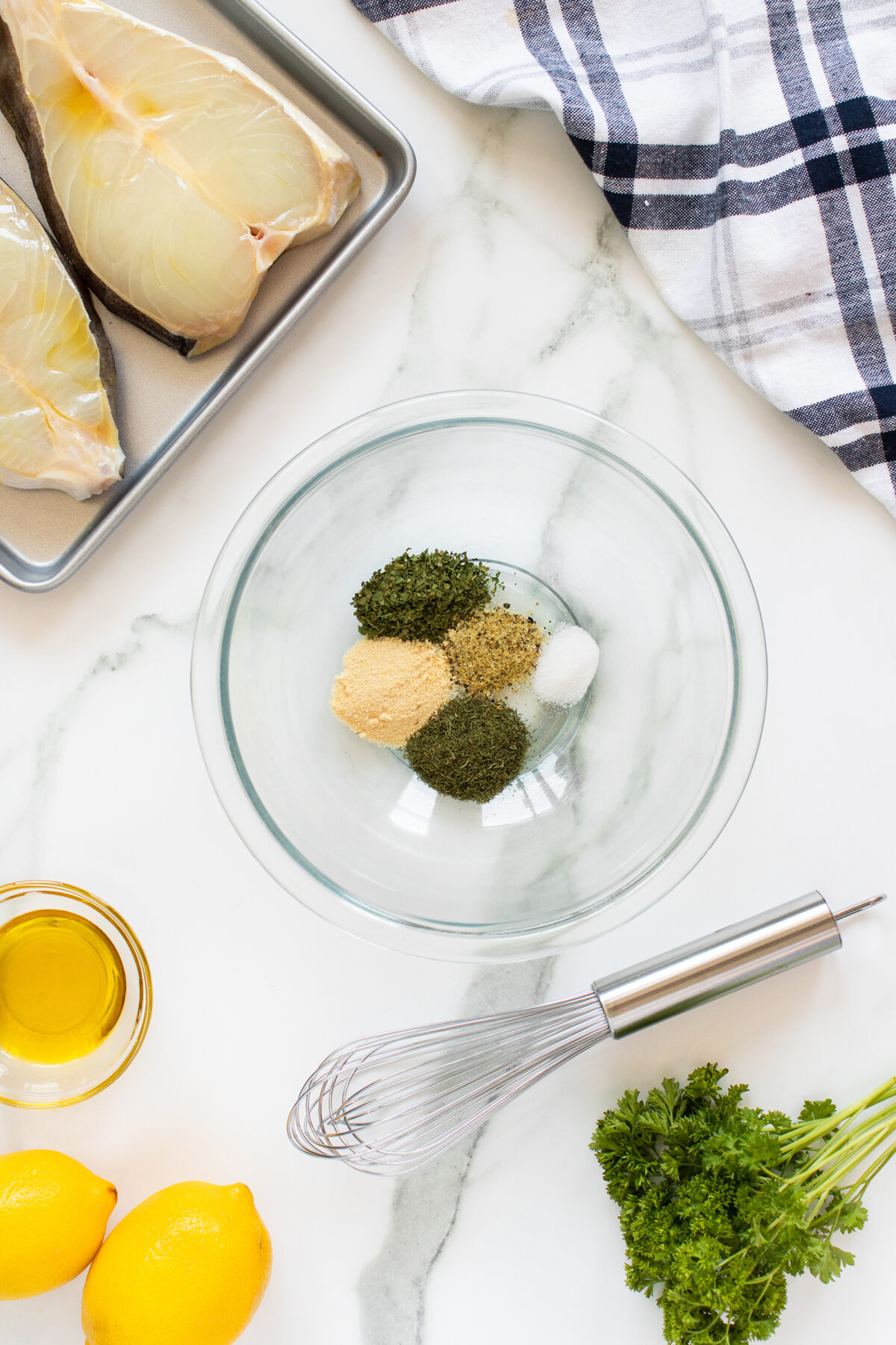 A glass bowl with assorted dried herbs and spices on a marble surface, next to a metal tray with fish fillets, a whisk, olive oil, lemons, fresh parsley, and a folded checkered dish towel.