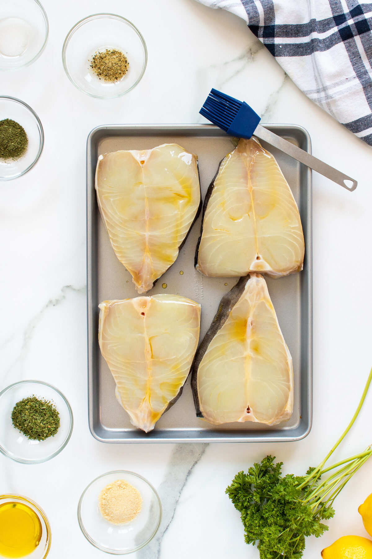 Four raw fish steaks are arranged on a metal baking tray with oil on top. Surrounding the tray are small bowls of seasonings, a brush, parsley, olive oil, and a striped kitchen towel on a marble surface.