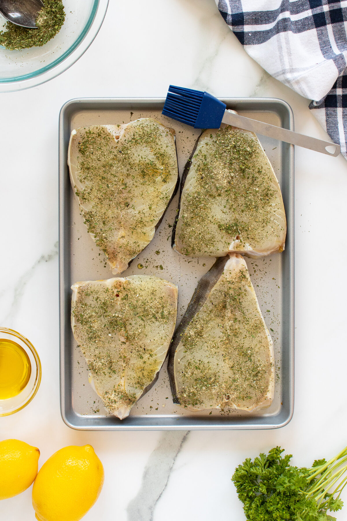 Four raw fish steaks seasoned with herbs are arranged on a metal baking tray. A blue basting brush rests on the tray. Nearby are a bowl, a towel, parsley, two lemons, and a small jar of olive oil.