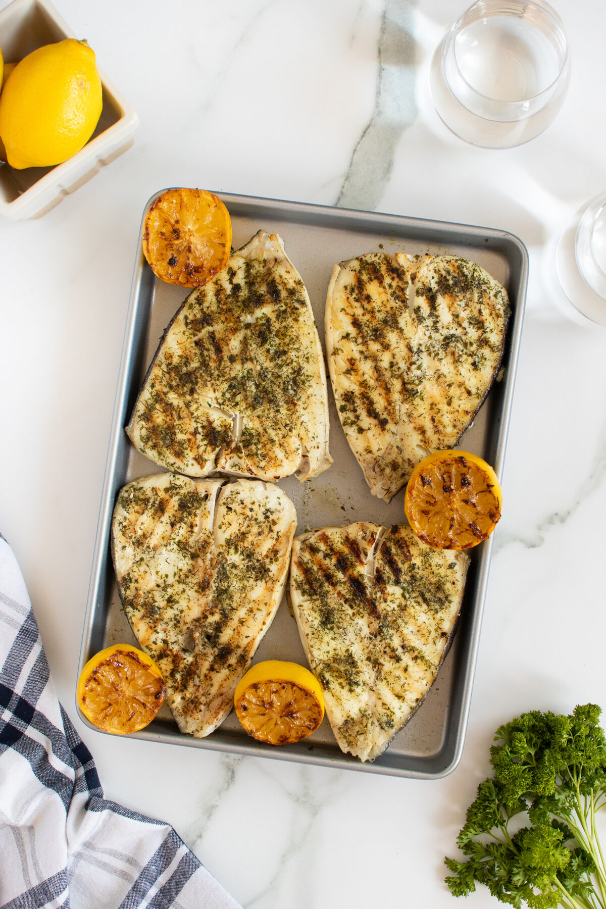 A baking sheet with four grilled swordfish steaks, seasoned with herbs, garnished with grilled lemon halves. A fresh lemon, two glasses of water, and parsley are nearby on a white marble surface. A striped towel is in the corner.