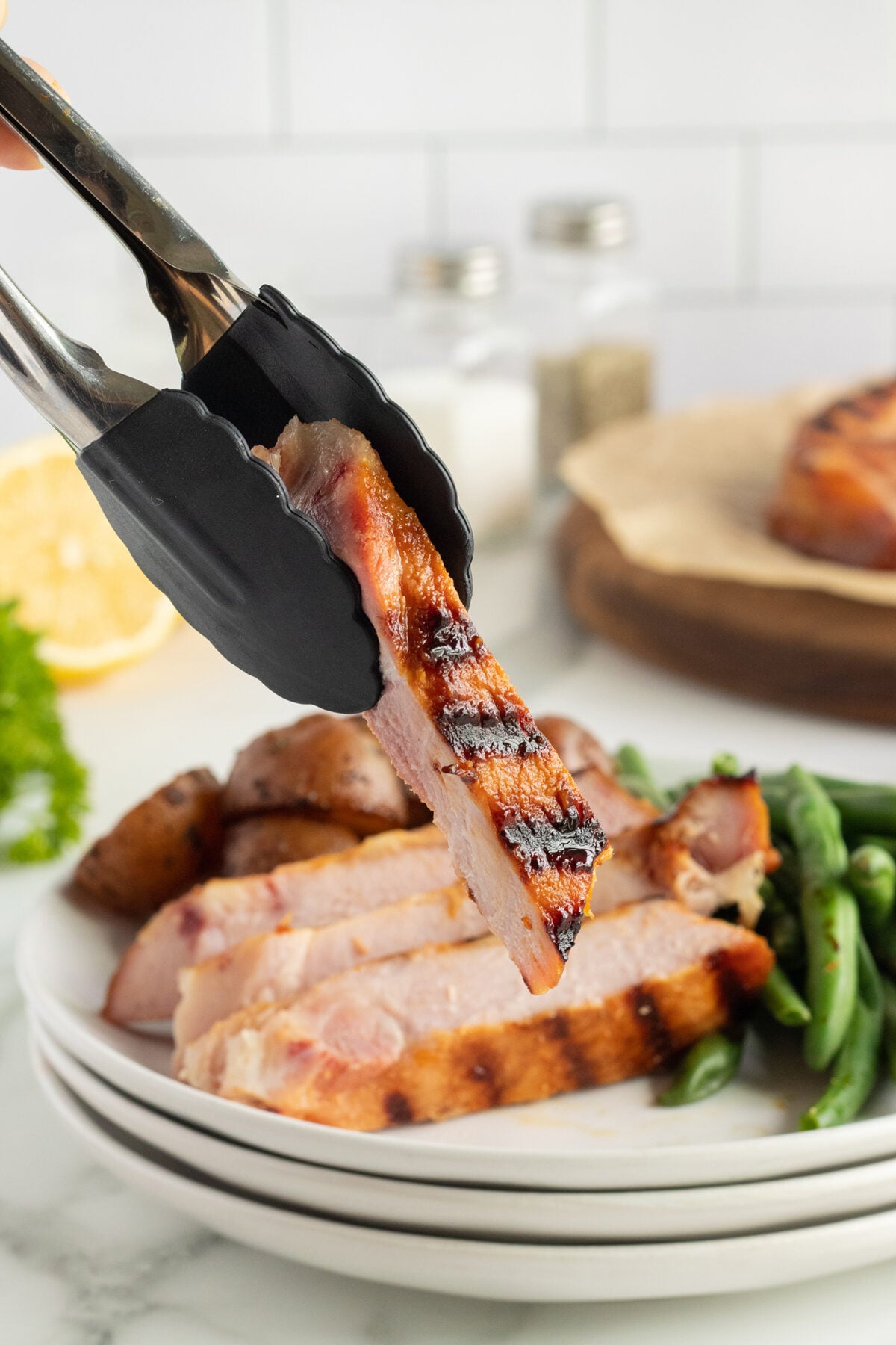 A pair of tongs holds a grilled pork chop slice above a plate with more pork slices, roasted potatoes, and green beans. Salt and pepper shakers are visible in the background.