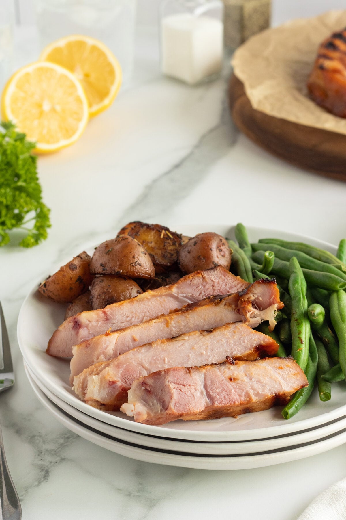 A plate of sliced roasted pork, green beans, and roasted potatoes sits on a marble surface. In the background are lemon halves, parsley, and a separate dish with more meat.