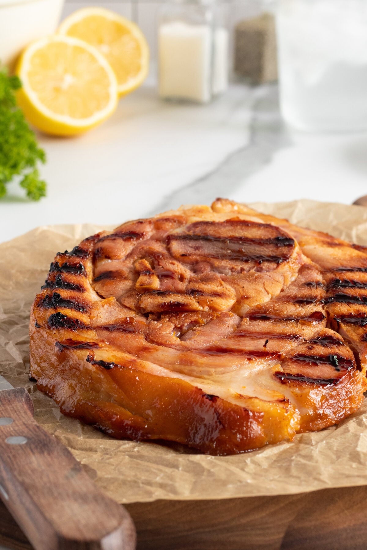 A grilled ham steak with caramelized edges sits on parchment paper, with a knife beside it. In the background are lemon halves, parsley, and salt and pepper shakers on a marble countertop.