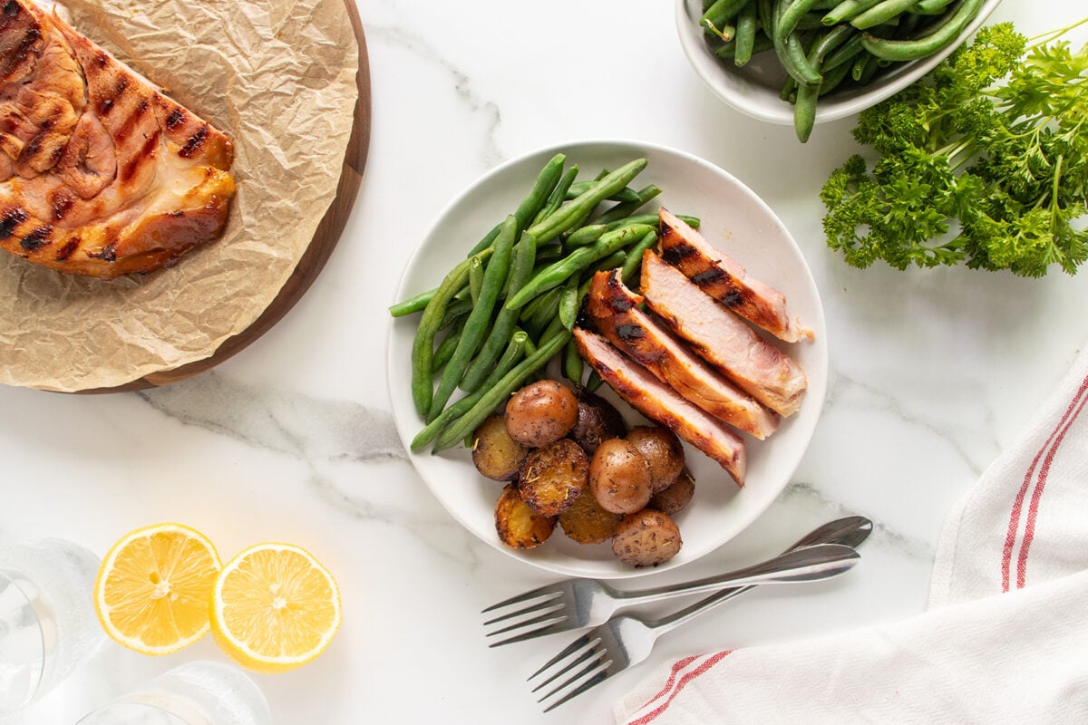 A plate with grilled ham slices, roasted baby potatoes, and green beans. Surrounding items include more green beans in a bowl, parsley, lemon halves, and grilled ham on parchment. Two forks rest by the plate.