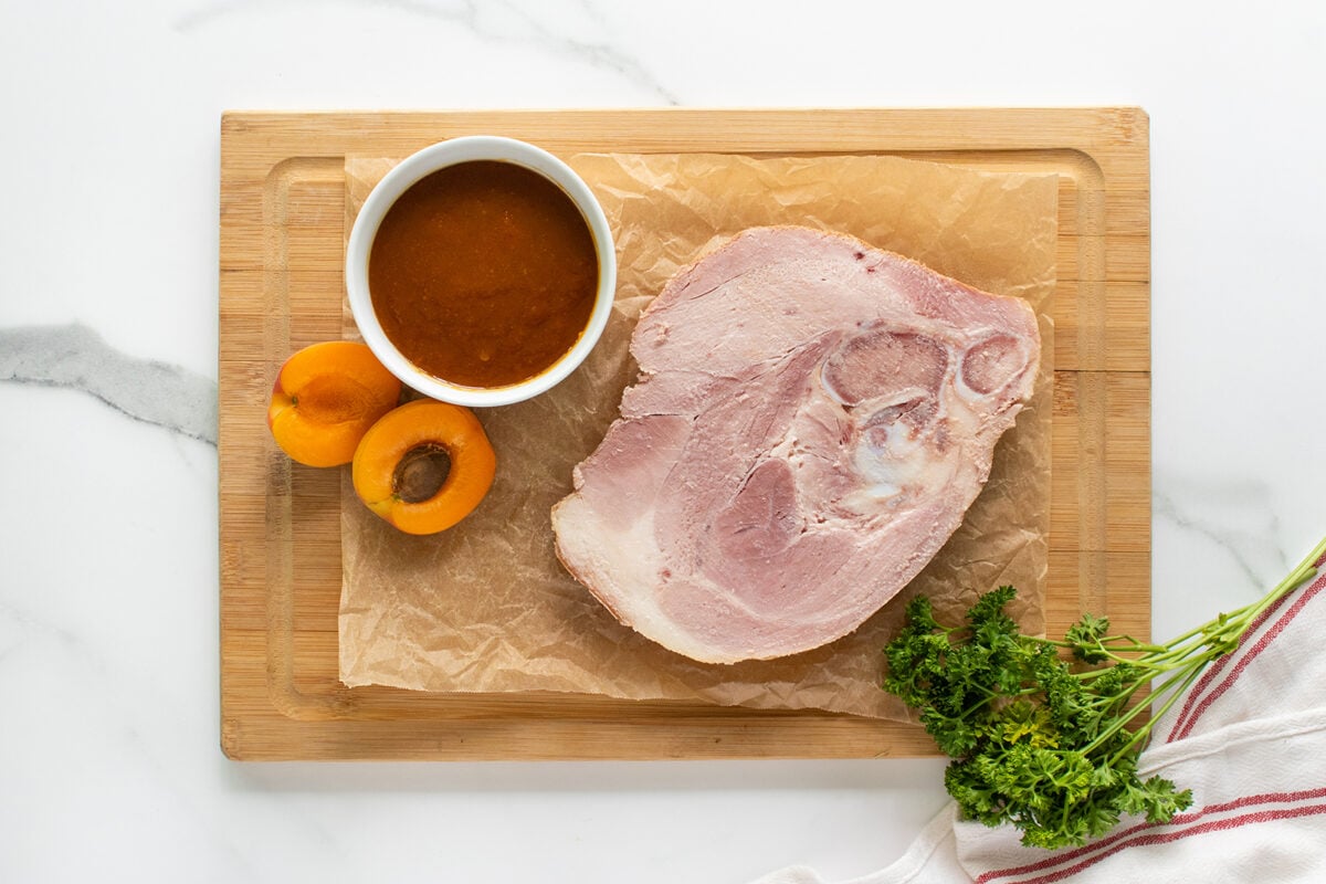 A wooden cutting board with a large slice of ham, a bowl of sauce, two halved apricots, and a bunch of fresh parsley on parchment paper, set on a white countertop with a striped towel nearby.
