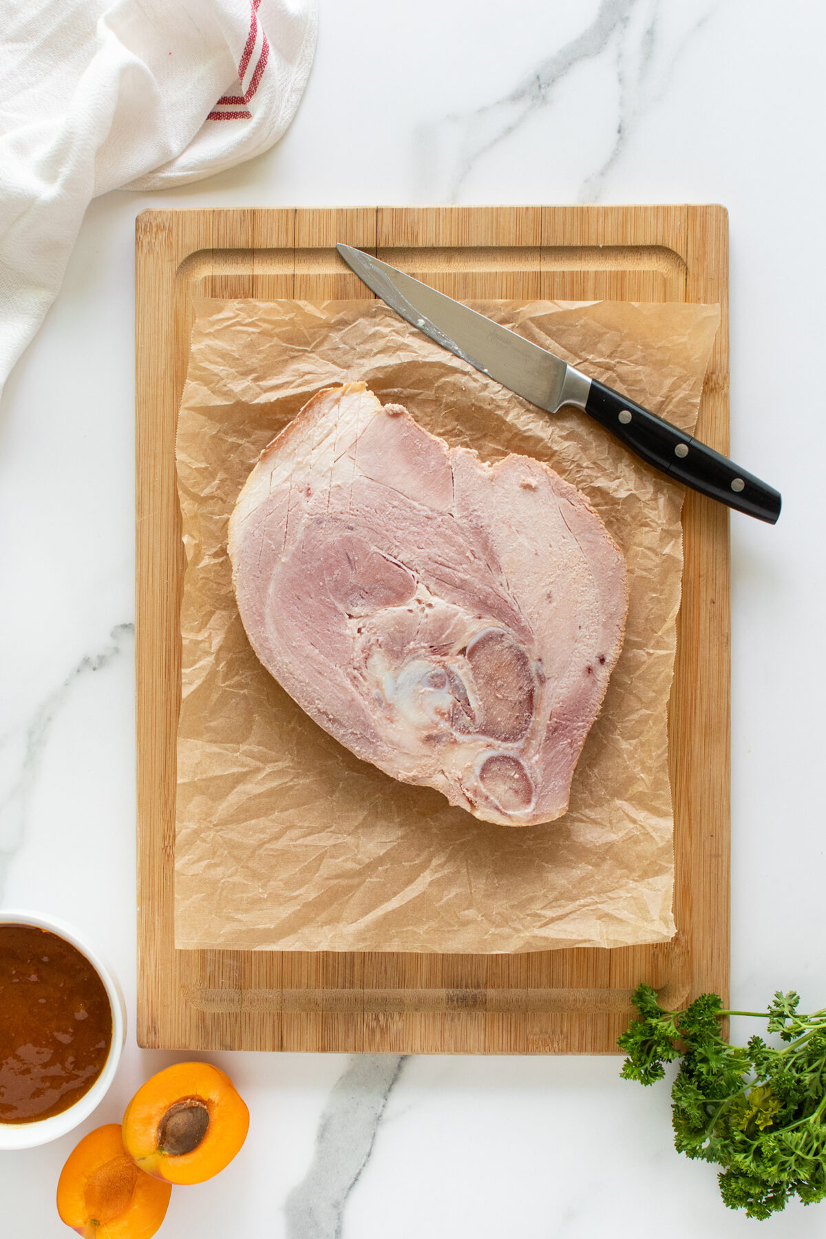 A raw ham steak sits on brown parchment paper on a wooden cutting board, with a knife beside it. Nearby are a bowl of sauce, a halved apricot, parsley, and a white cloth on a marble surface.