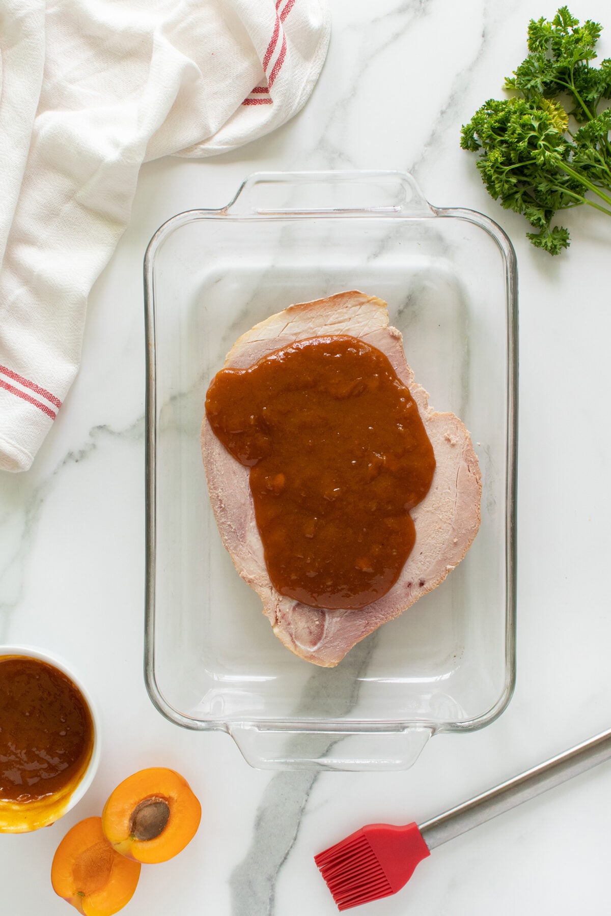 A glass baking dish with a ham slice topped with brown apricot glaze. Nearby are a halved apricot, a bowl of glaze, a red basting brush, parsley, and a white towel on a marble surface.