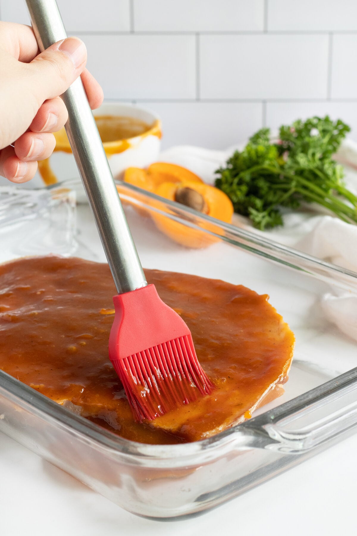 A hand uses a red silicone brush to spread sauce over a round piece of food in a glass baking dish. Fresh herbs and a halved apricot are visible in the background on a white surface.