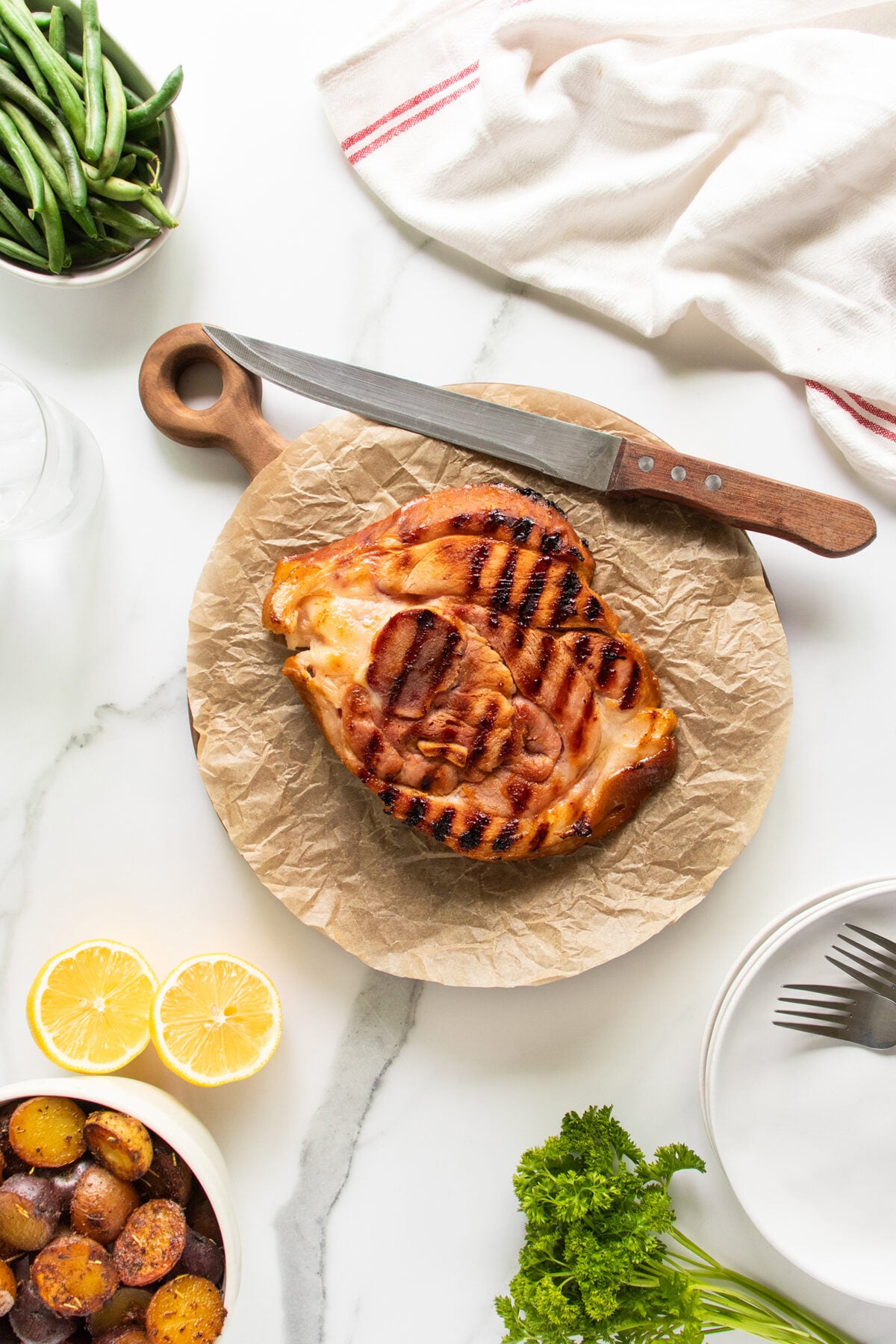 Grilled steak with char marks on parchment paper atop a wooden board, beside a knife. Surrounding items include green beans, lemon halves, roasted potatoes, parsley, a white napkin, and empty plates on a white surface.