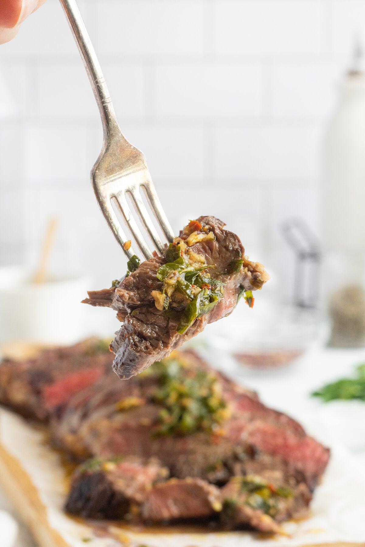 A fork holds a piece of medium-rare steak topped with chimichurri sauce; the rest of the sliced steak and sauce are blurred in the background on a white surface.
