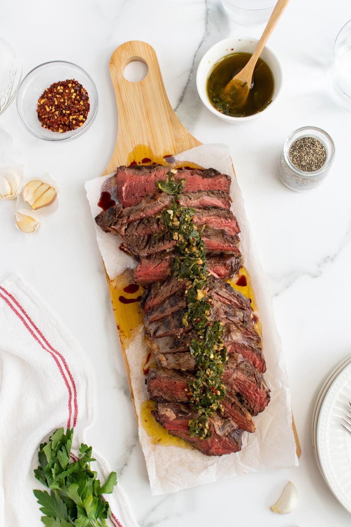 Sliced grilled steak topped with chimichurri sauce on a wooden cutting board, surrounded by garlic, dried chili flakes, parsley, pepper, and a towel on a white marble surface.