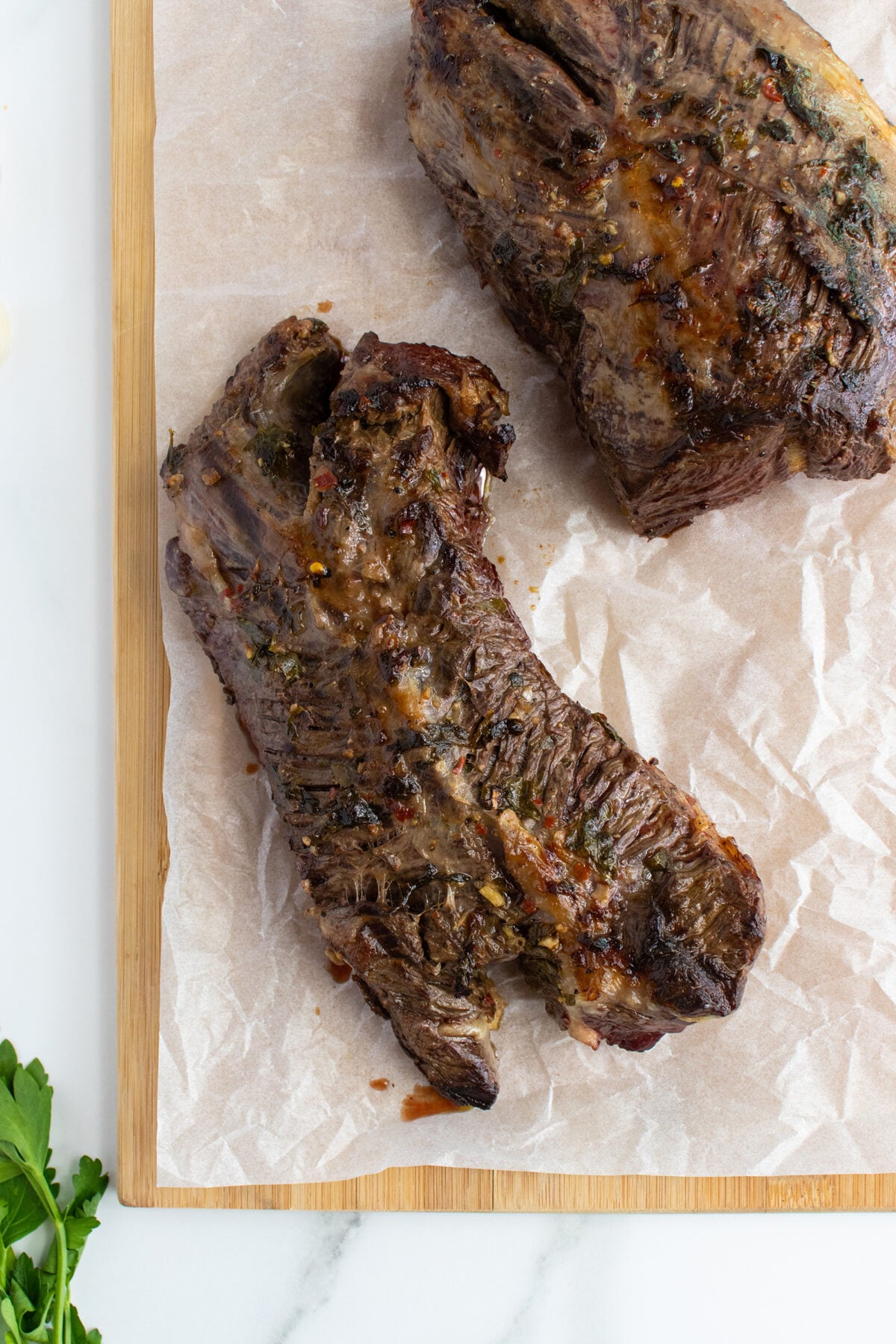 Two pieces of cooked beef short ribs resting on parchment paper atop a wooden cutting board, with a sprig of parsley visible in the lower left corner.