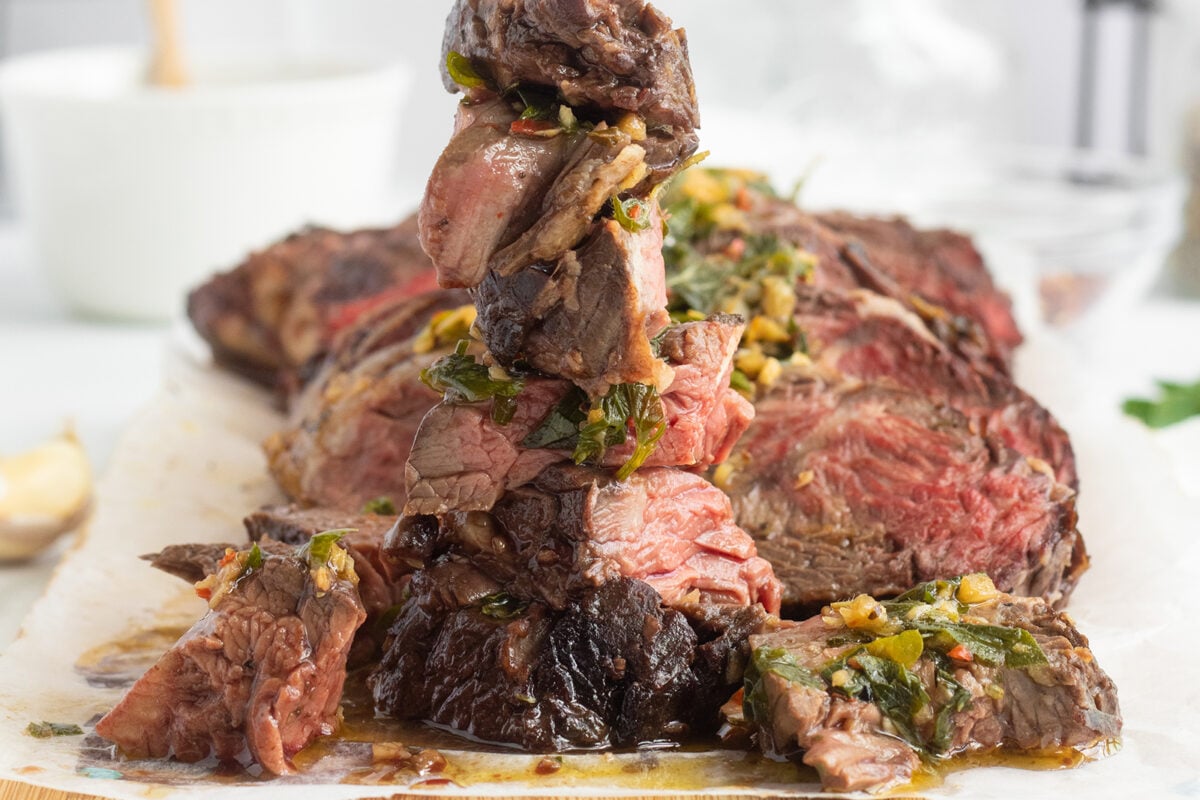 Sliced medium-rare steak stacked vertically, topped with a fresh herb and garlic sauce, displayed on parchment paper with more steak pieces in the background.