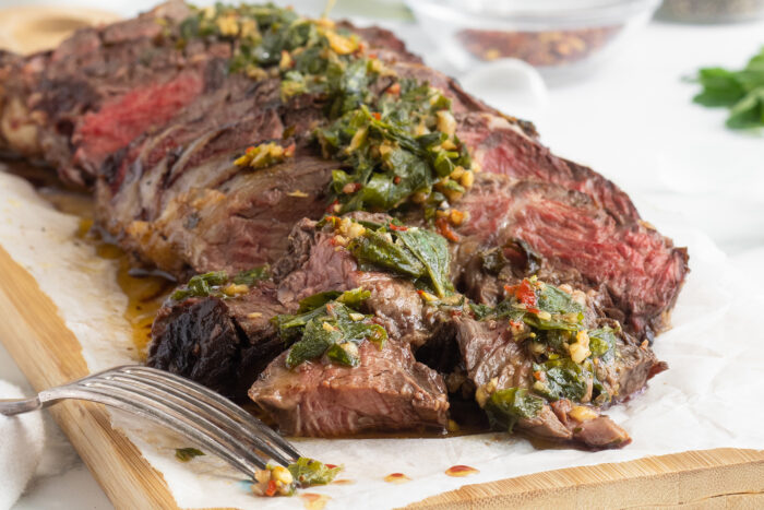 Sliced medium-rare steak topped with a green herb sauce, served on parchment paper on a wooden board, with a fork beside the meat. Fresh herbs and a glass bowl are blurred in the background.