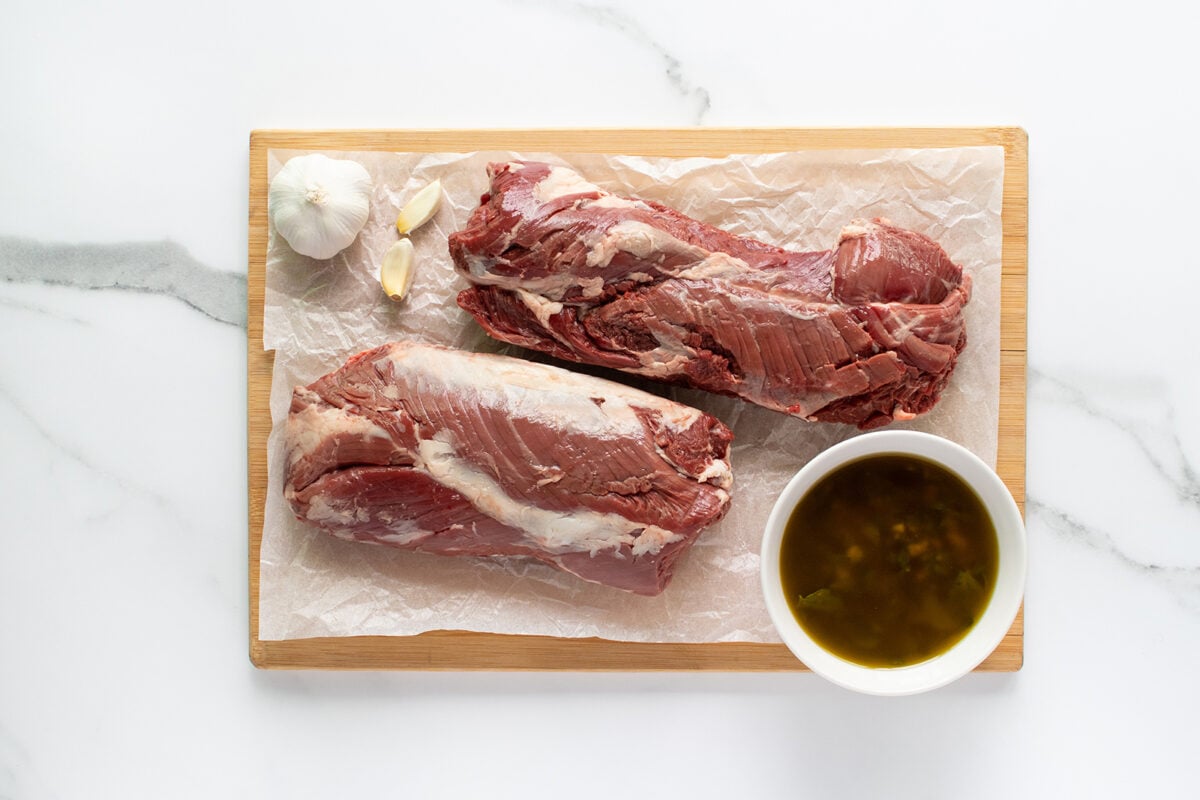 Two raw pieces of beef on a parchment-lined wooden cutting board, with a whole garlic bulb, garlic cloves, and a small bowl of marinade or sauce next to the meat, all on a white marble surface.