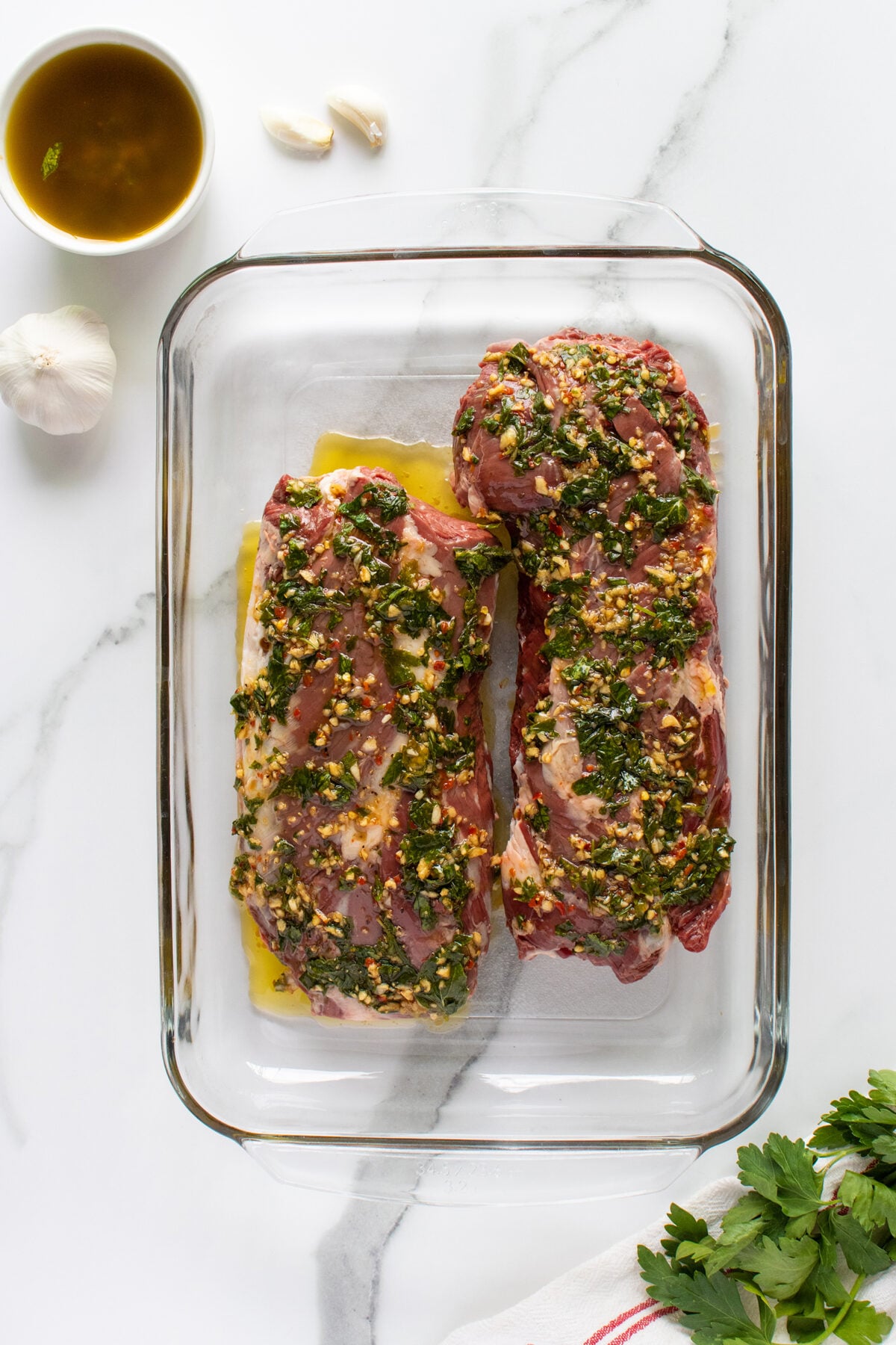 Two raw pork tenderloins coated with a herb and garlic marinade in a glass baking dish, placed on a white marble surface with a bowl of oil, whole garlic, and fresh parsley nearby.