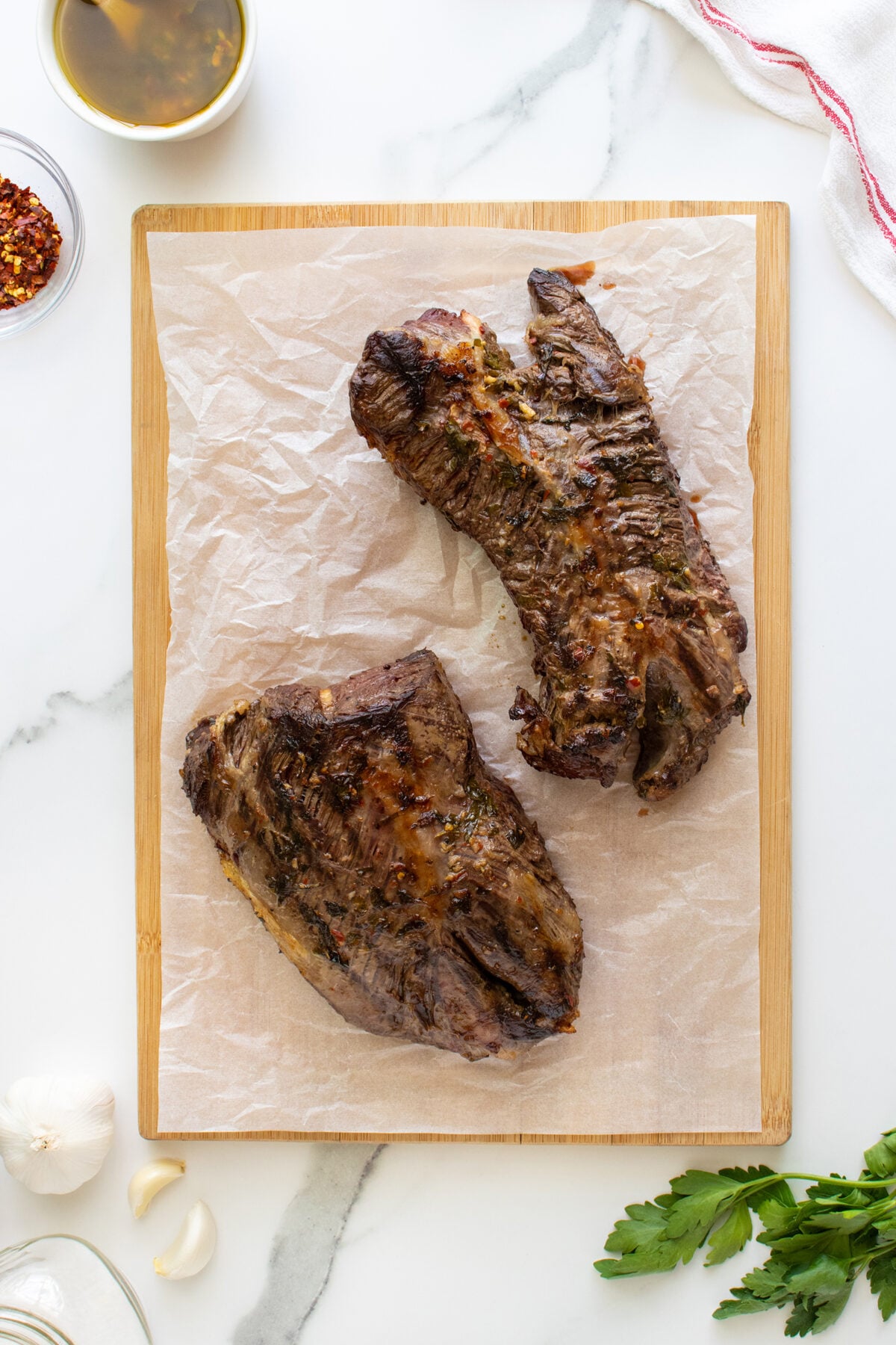 Two grilled steak pieces resting on parchment paper atop a wooden cutting board, surrounded by parsley, garlic, chili flakes, and a bowl of oil on a white marble surface.