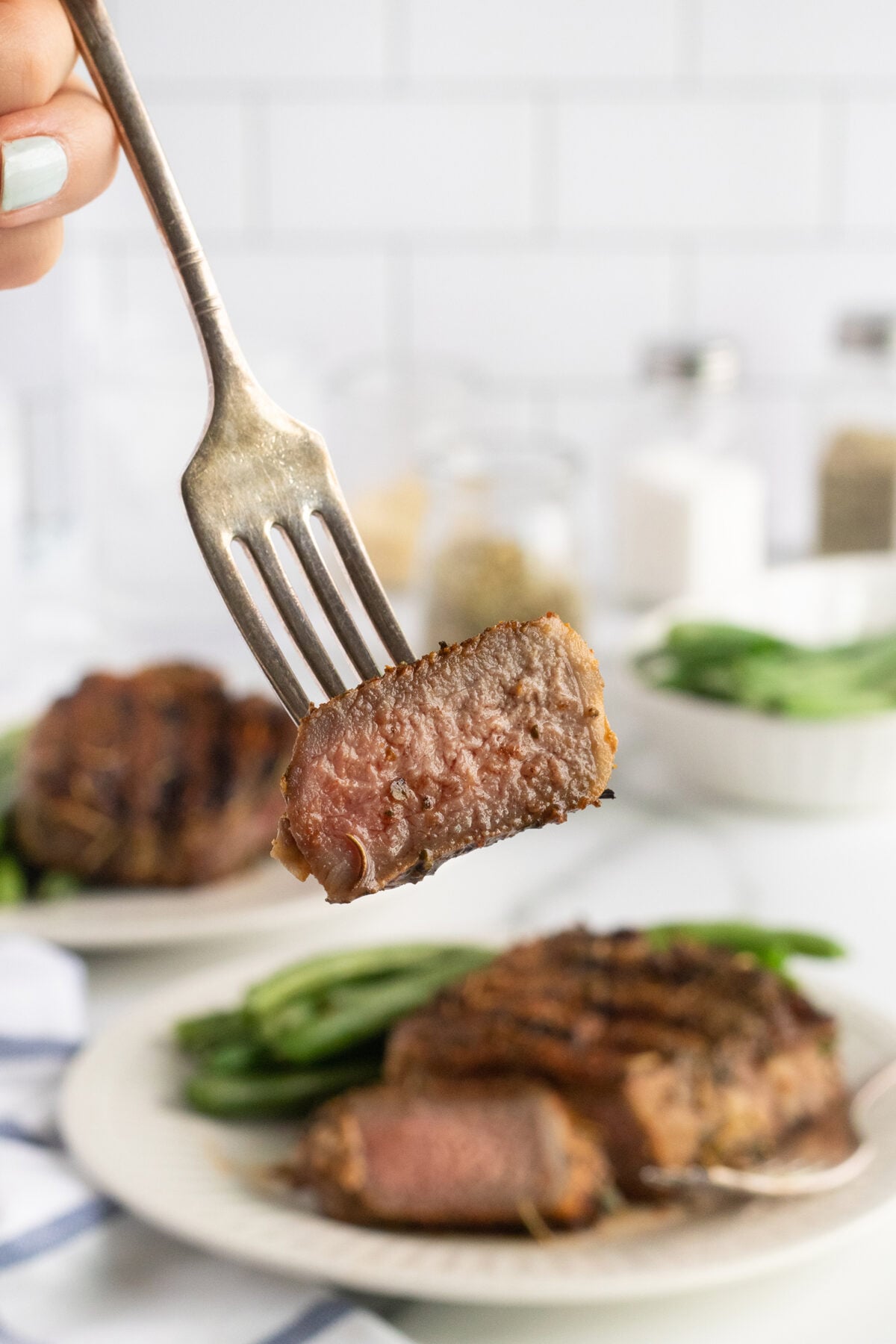 A close-up of a fork holding a piece of medium-rare steak above a plate with more steak and green beans, with a blurred kitchen background.