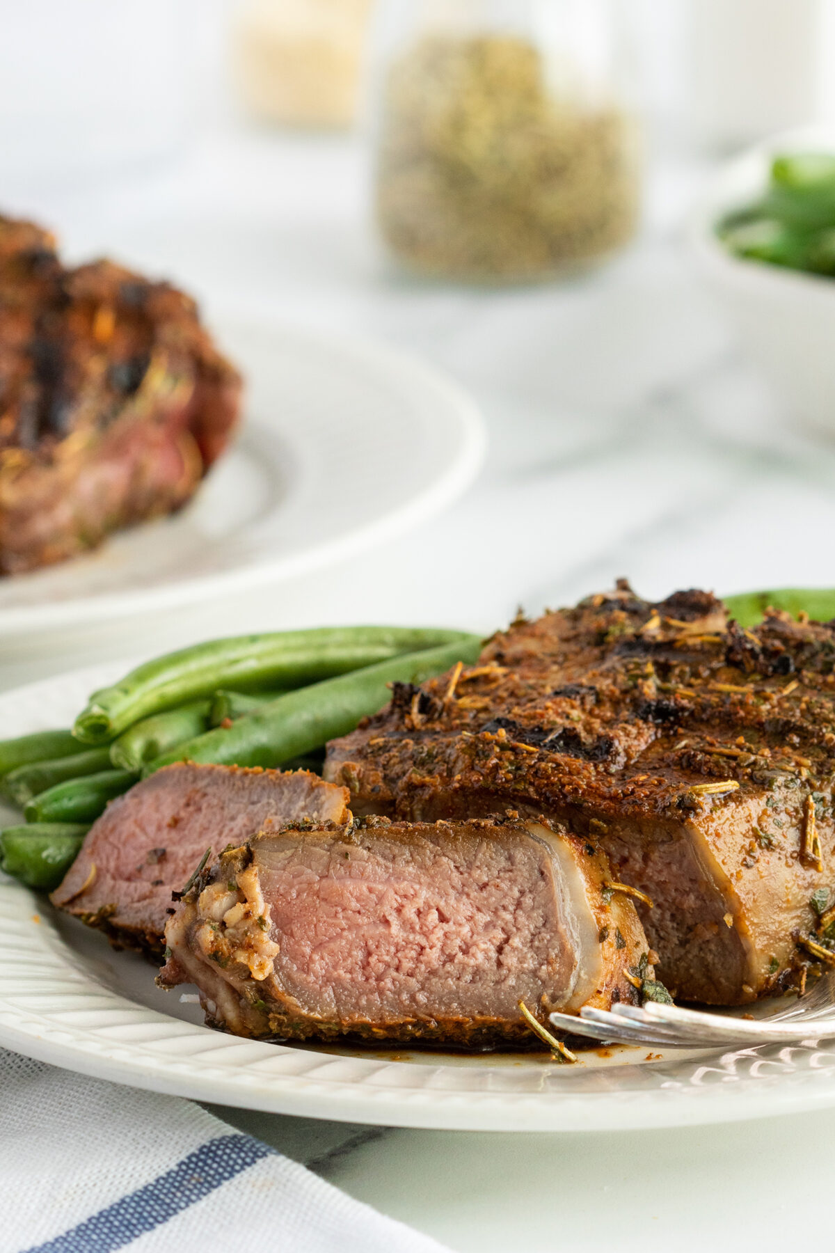 A plate with a sliced, seasoned steak cooked medium-rare, served with fresh green beans. A fork rests on the plate, and another steak is blurred in the background.