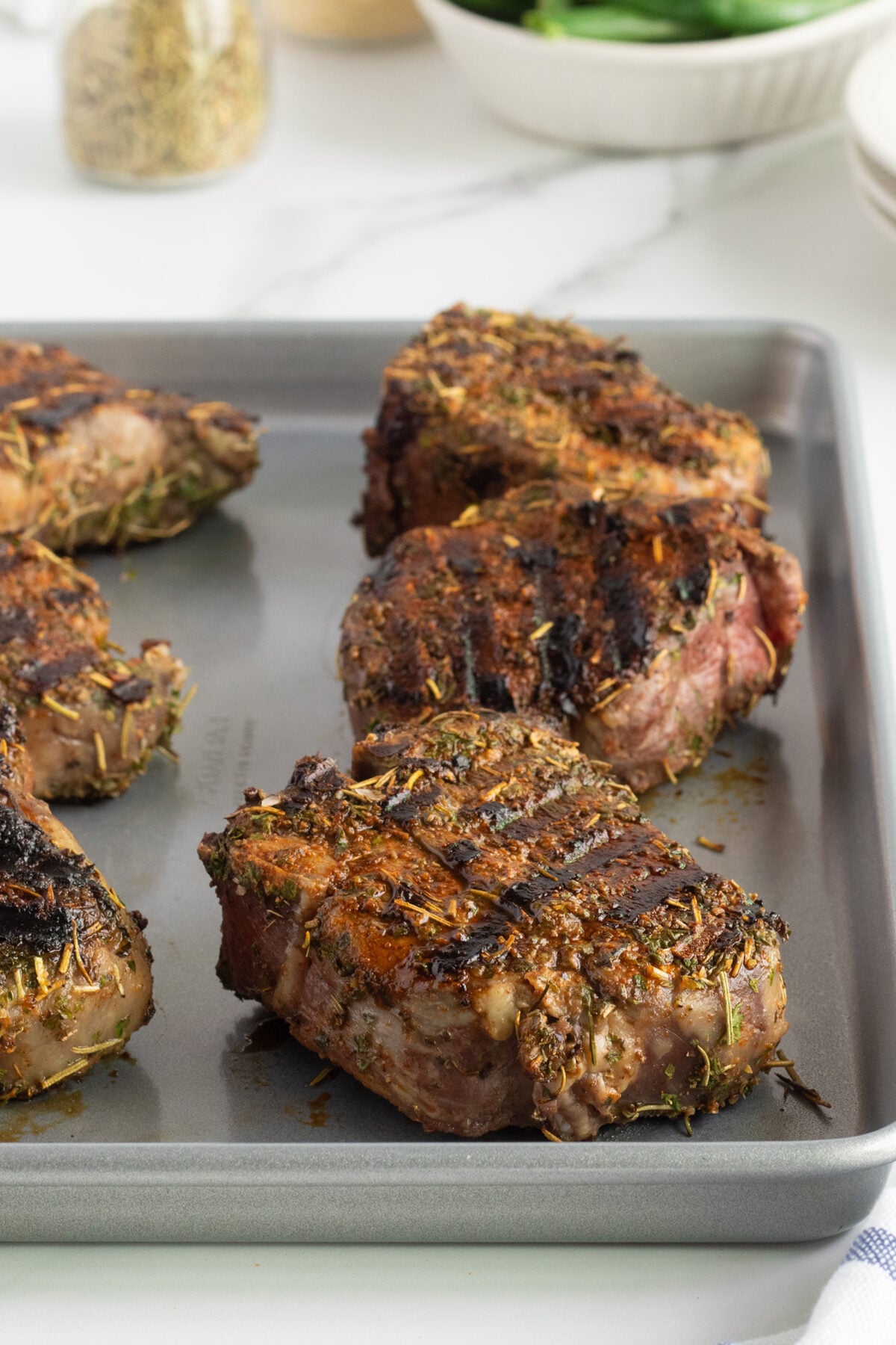 Four grilled lamb chops with herb seasoning rest on a metal baking sheet. The meat shows char marks and a browned crust, with some herbs visible on the surface. A bowl and spice jar are blurred in the background.