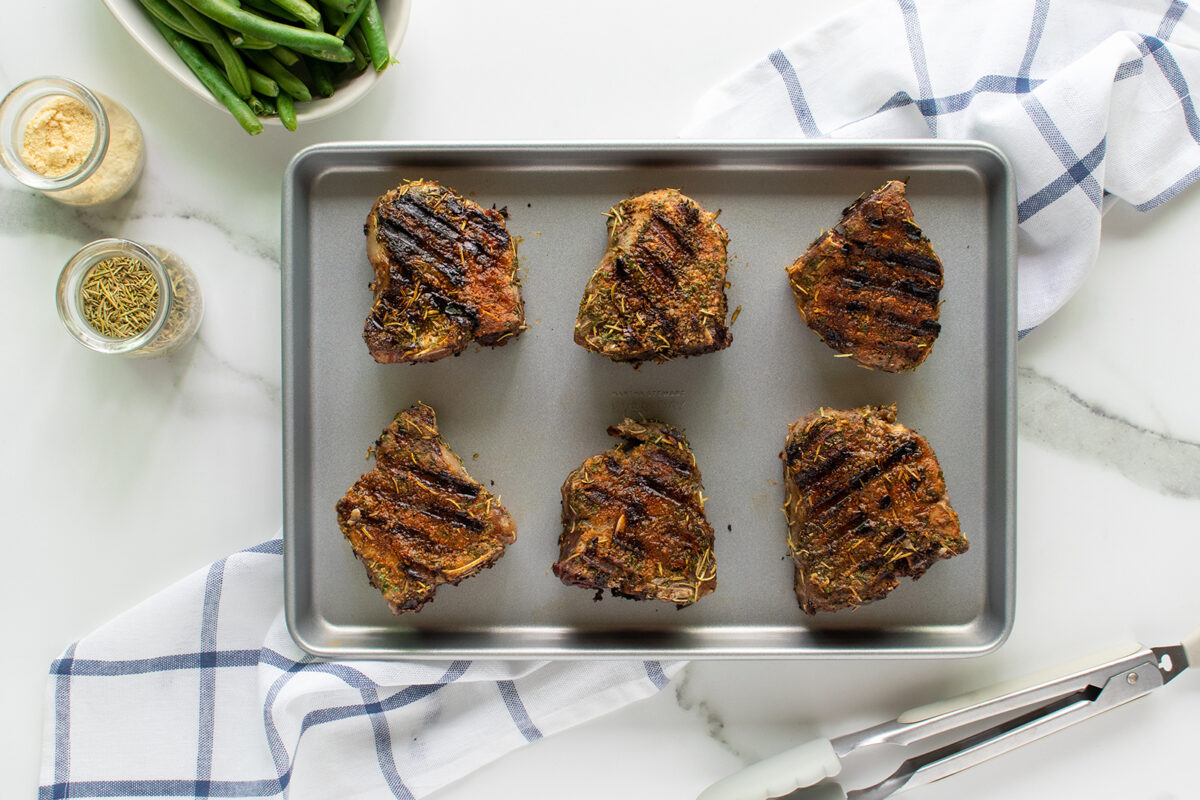 Six grilled lamb chops with visible grill marks on a baking tray, surrounded by a jar of spices, a bowl of green beans, kitchen tongs, and a blue-and-white checkered towel on a white countertop.