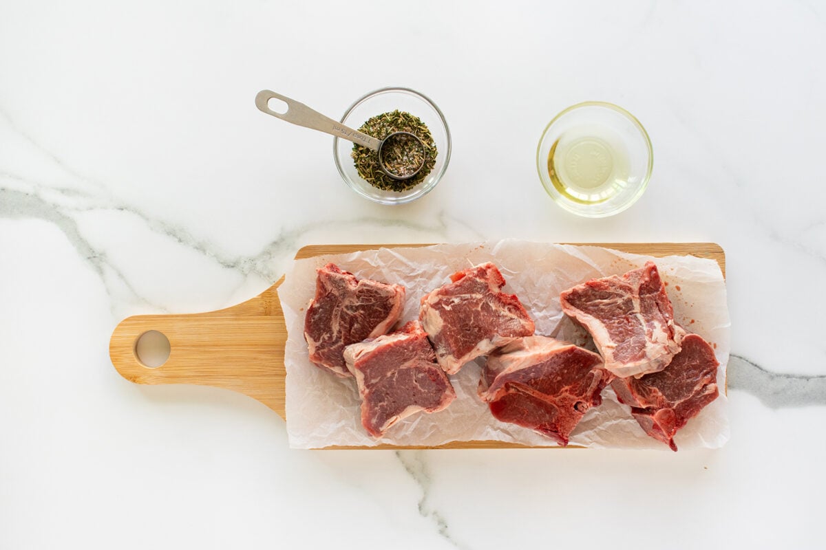 Raw lamb chops are arranged on a wooden cutting board lined with parchment paper. Above the board, there is a small bowl of dried herbs with a measuring spoon and a glass bowl containing olive oil.