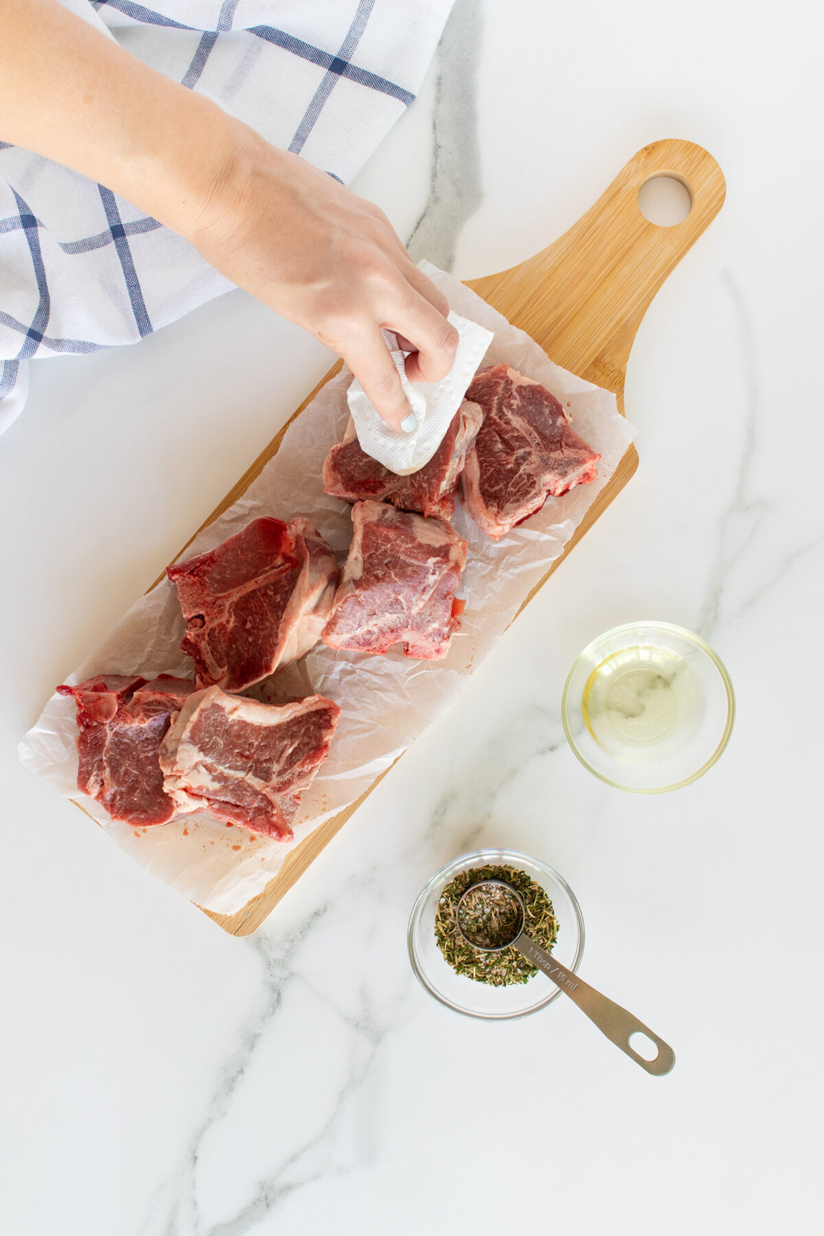 A hand uses a paper towel to pat dry raw beef pieces on parchment paper atop a wooden cutting board. Nearby are a small bowl of oil and a bowl of dried herbs with a measuring spoon. A blue-striped cloth is partially visible.