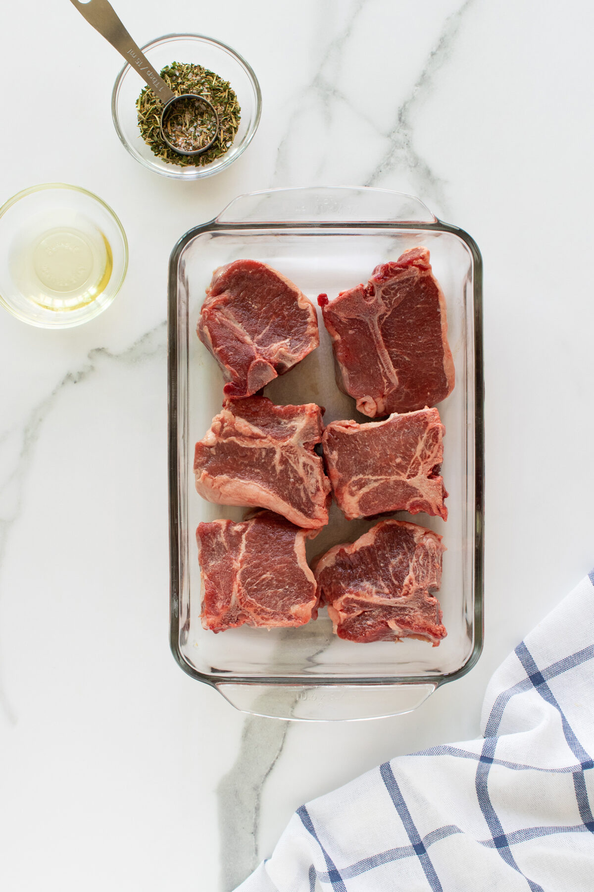 Six raw lamb chops in a glass baking dish on a marble surface, with a bowl of oil, a small bowl of dried herbs, a metal measuring spoon, and a blue-striped kitchen towel nearby.